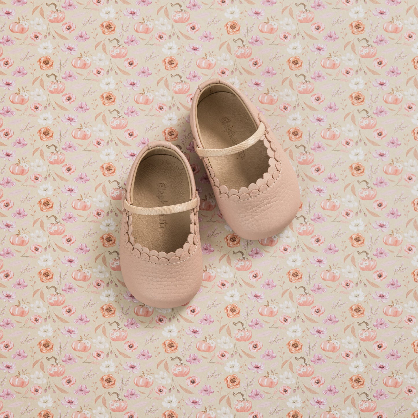 Soft pink baby Mary Jane shoes with scalloped edges and strap, on floral fabric background.