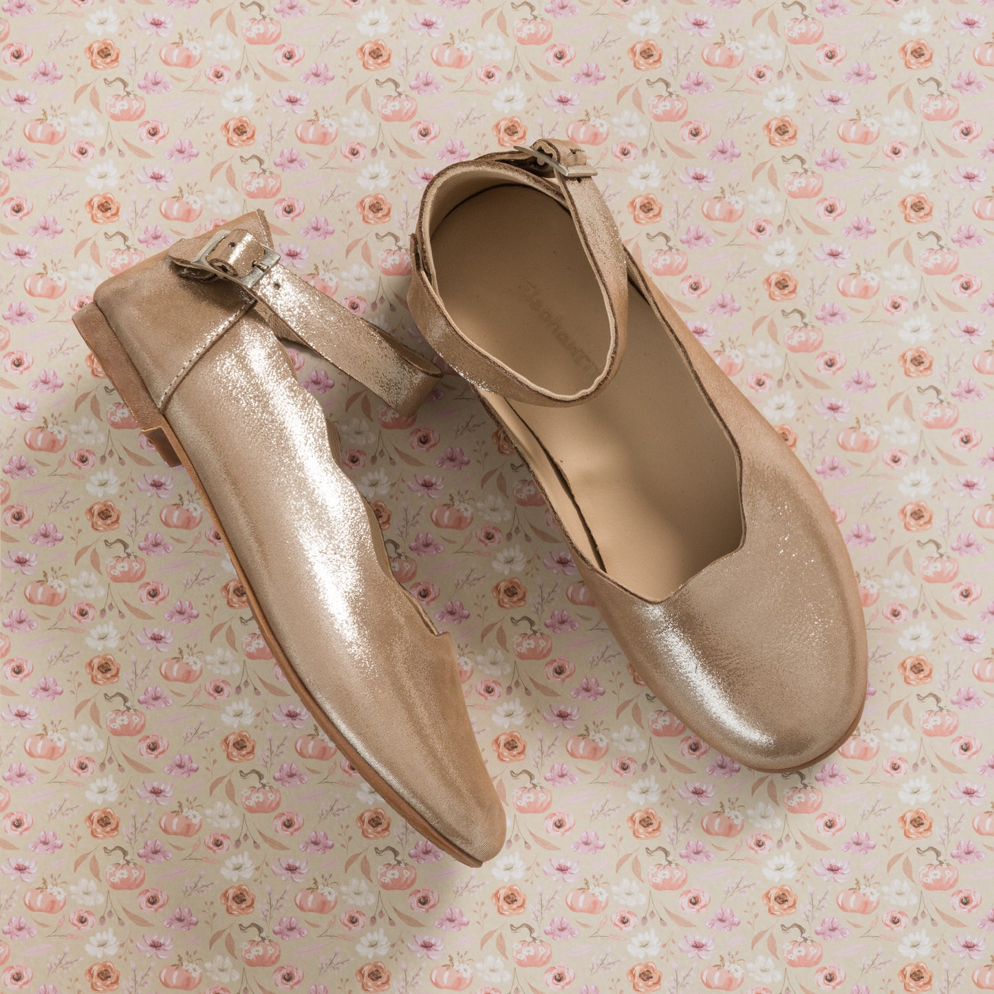 Blush pink toddler ballerina flats with ankle strap and subtle sheen on floral background.
