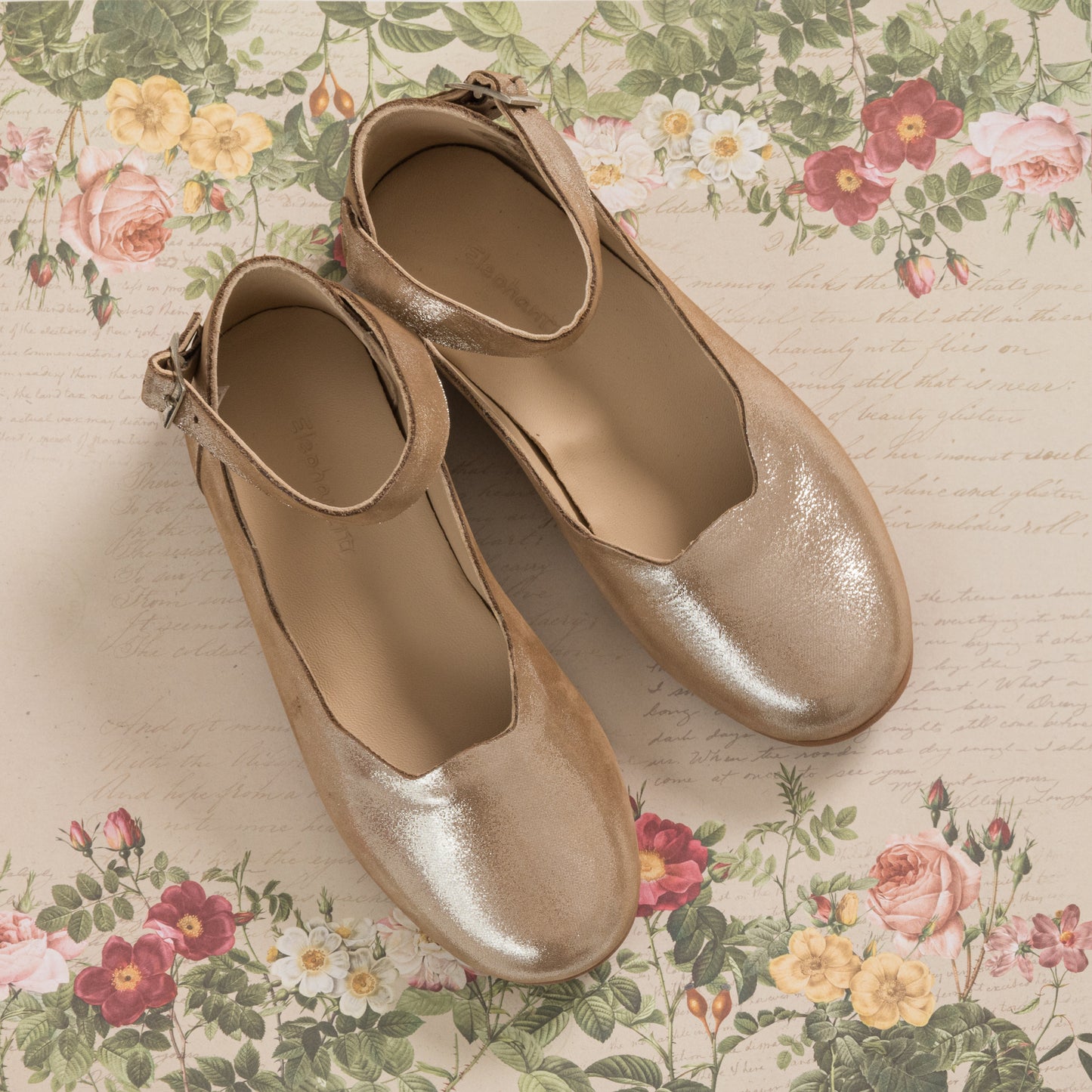 Shiny blush pink toddler flats with ankle straps and rounded toes on floral background.