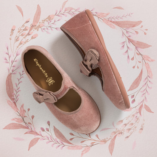 Pink velvet Mary Jane shoes with bow detail and rubber sole, displayed on floral background.