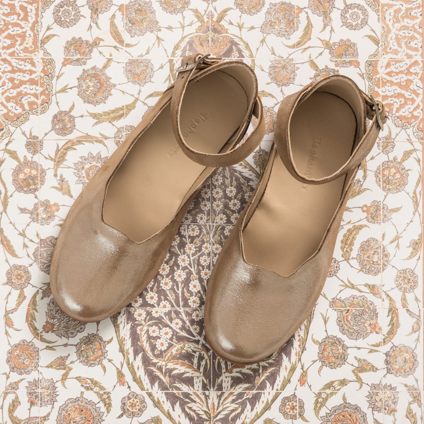 Blush pink toddler flats with ankle straps and rounded toes on floral fabric background.