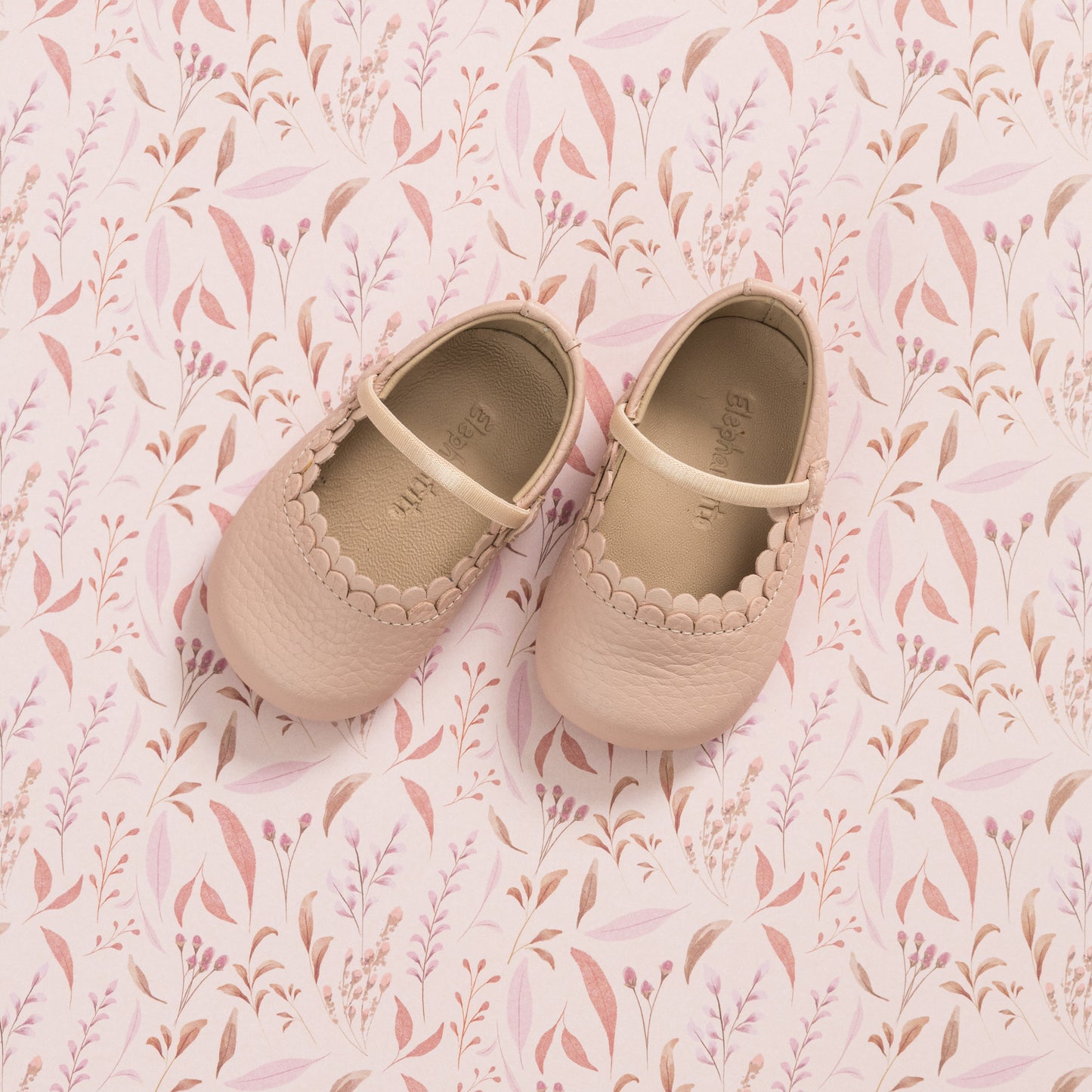 Soft pink baby shoes with scalloped edges and a Mary Jane strap on patterned pink background.