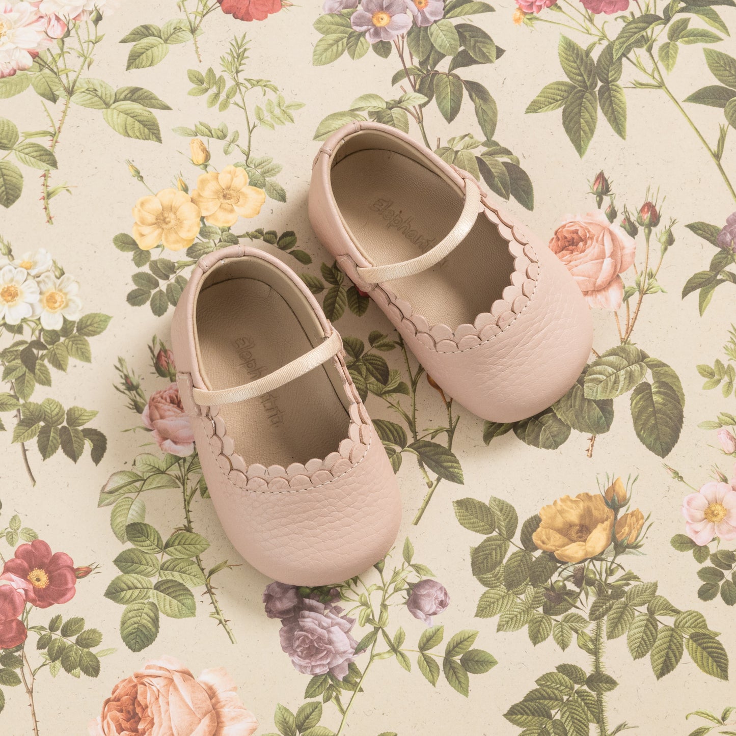 Soft pink baby Mary Jane shoes with scalloped edges and a single strap, resting on floral fabric background.