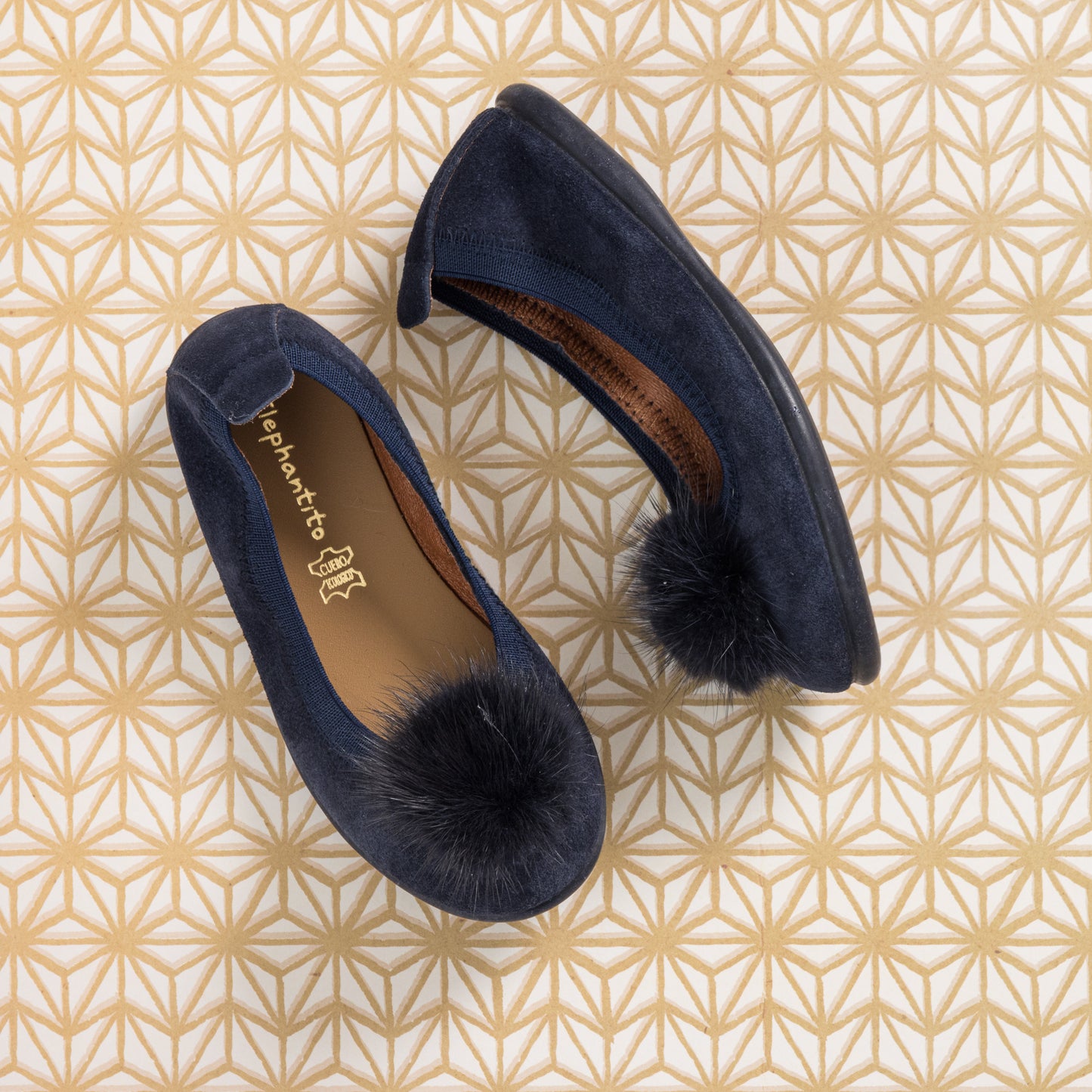 Blue suede ballerina flats with black pom poms on toes and brown insoles on geometric patterned background.