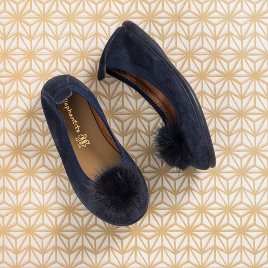 Blue suede ballerina flats with black pom poms on toes and brown insoles on geometric patterned background.
