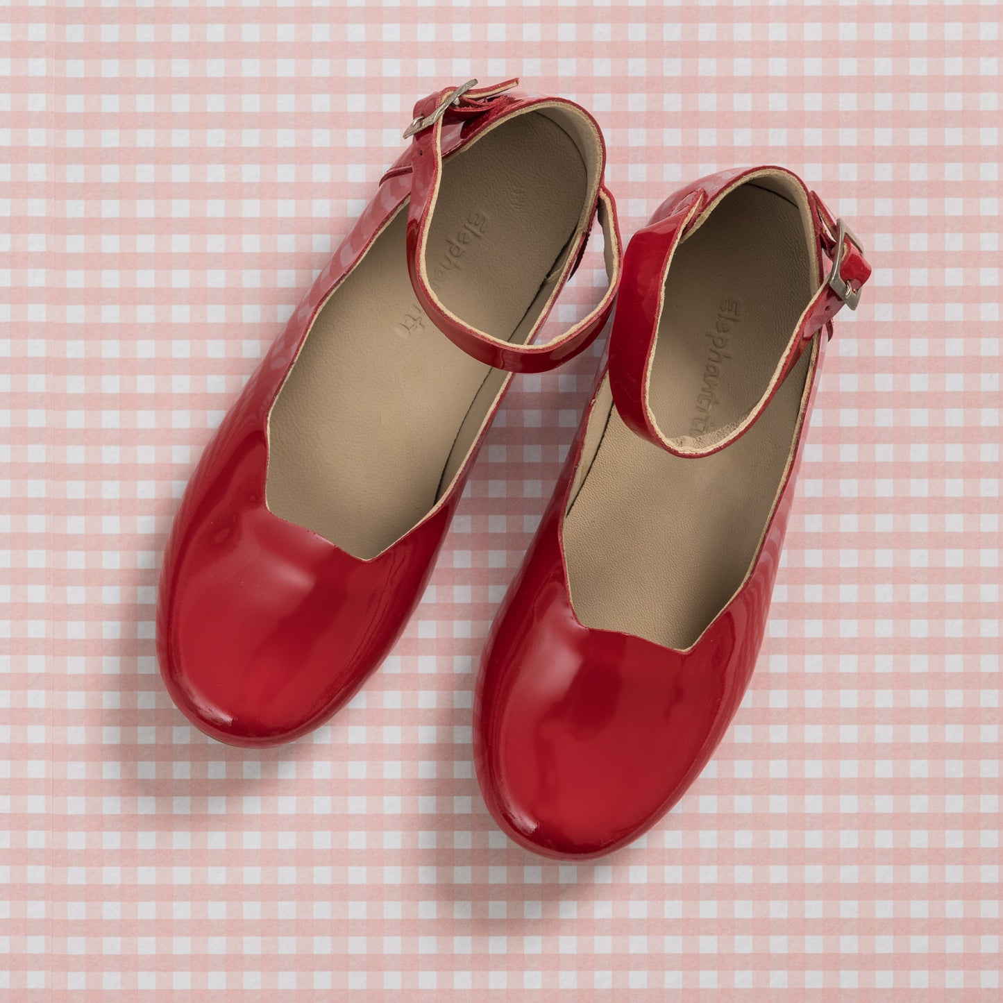Shiny red patent leather girls' ballerina flats with ankle straps on pink gingham background.