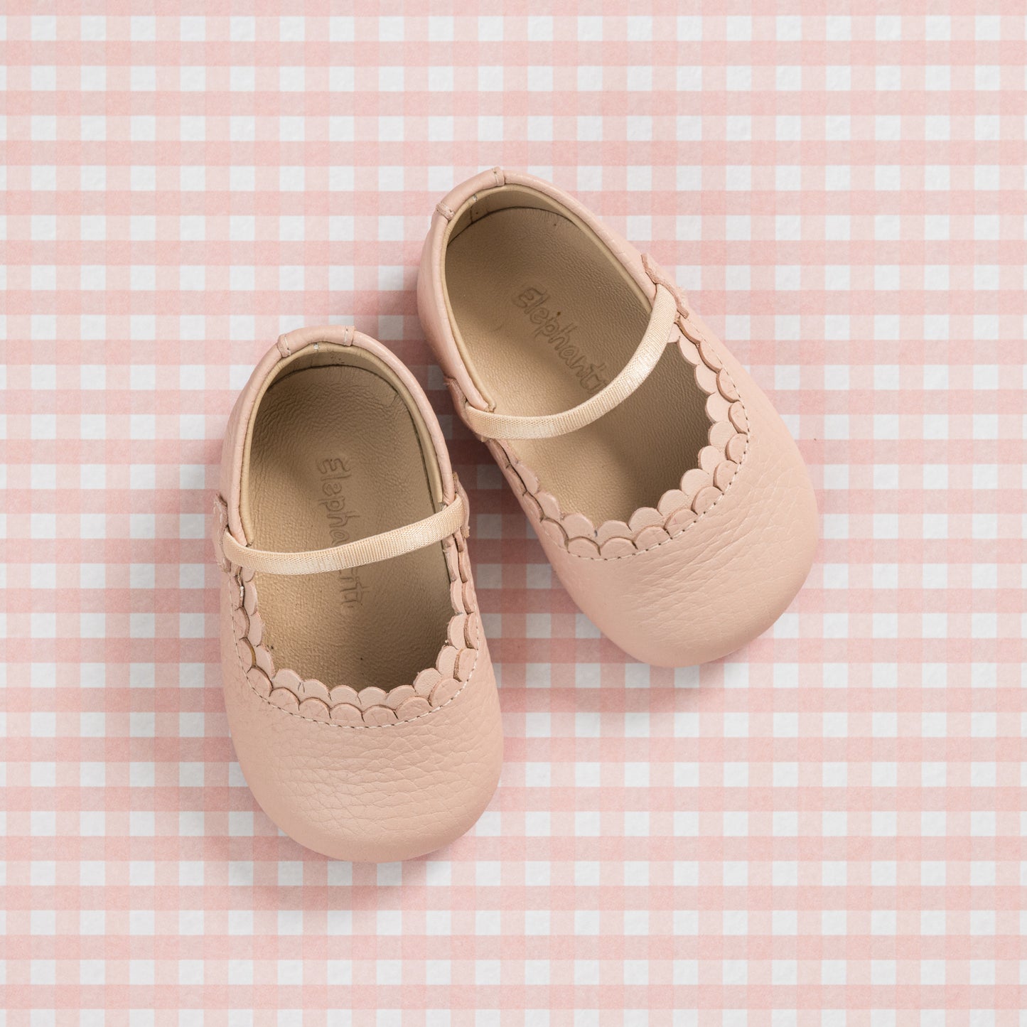 Soft pink baby shoes with scalloped edges and Mary Jane strap on gingham background.