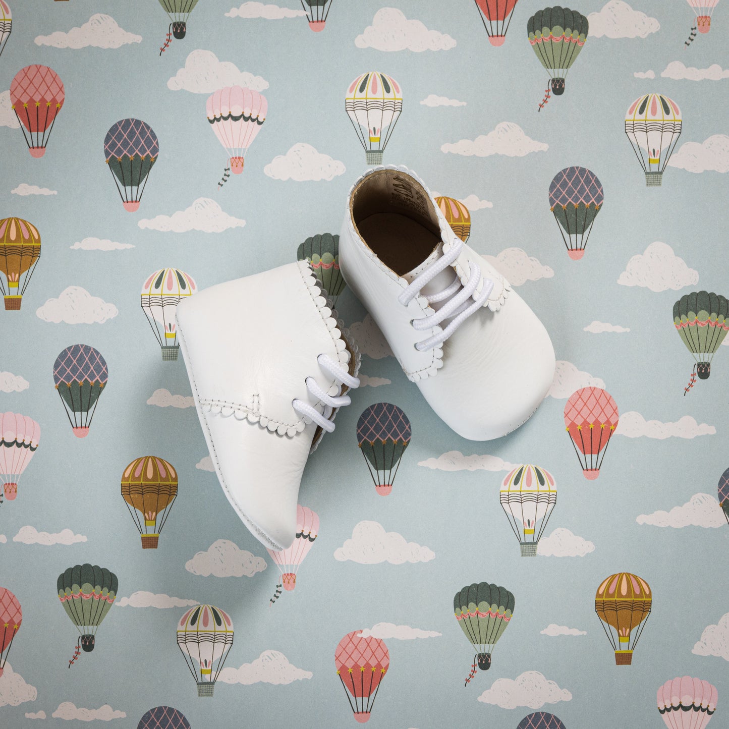 White baby booties with scalloped edges and white laces on pastel hot air balloon background.