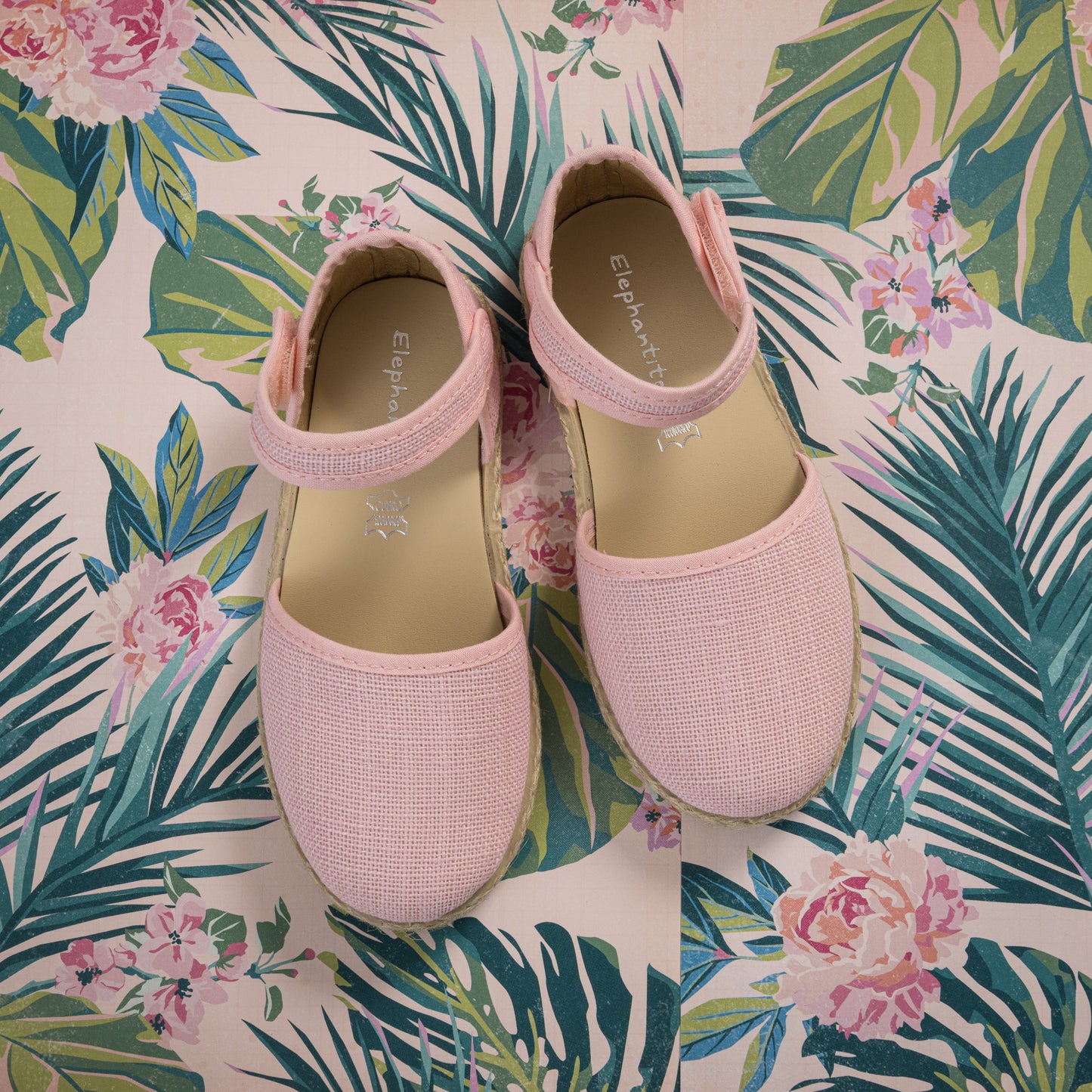 Pink linen espadrille shoes for toddlers with ankle straps and woven jute soles on floral background.