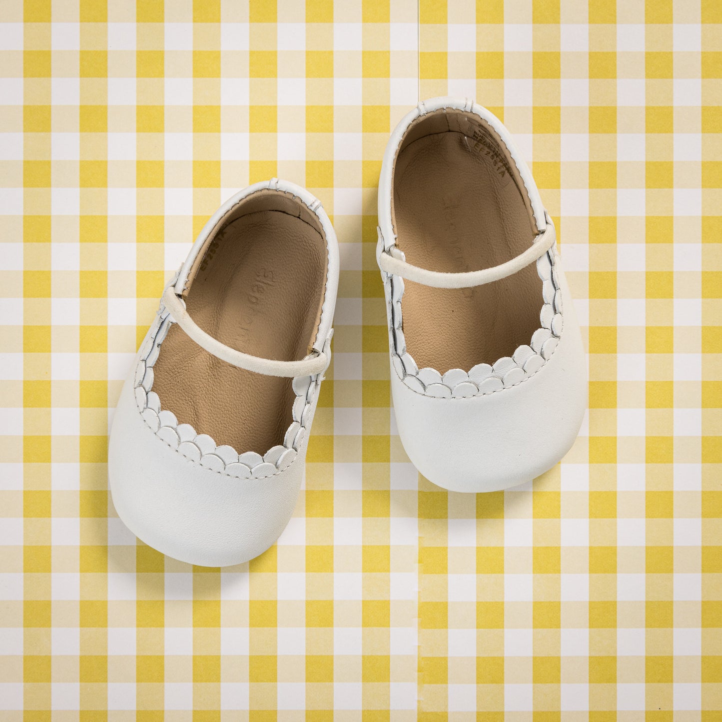 White baby Mary Jane shoes with scalloped edges and thin strap on yellow gingham background.