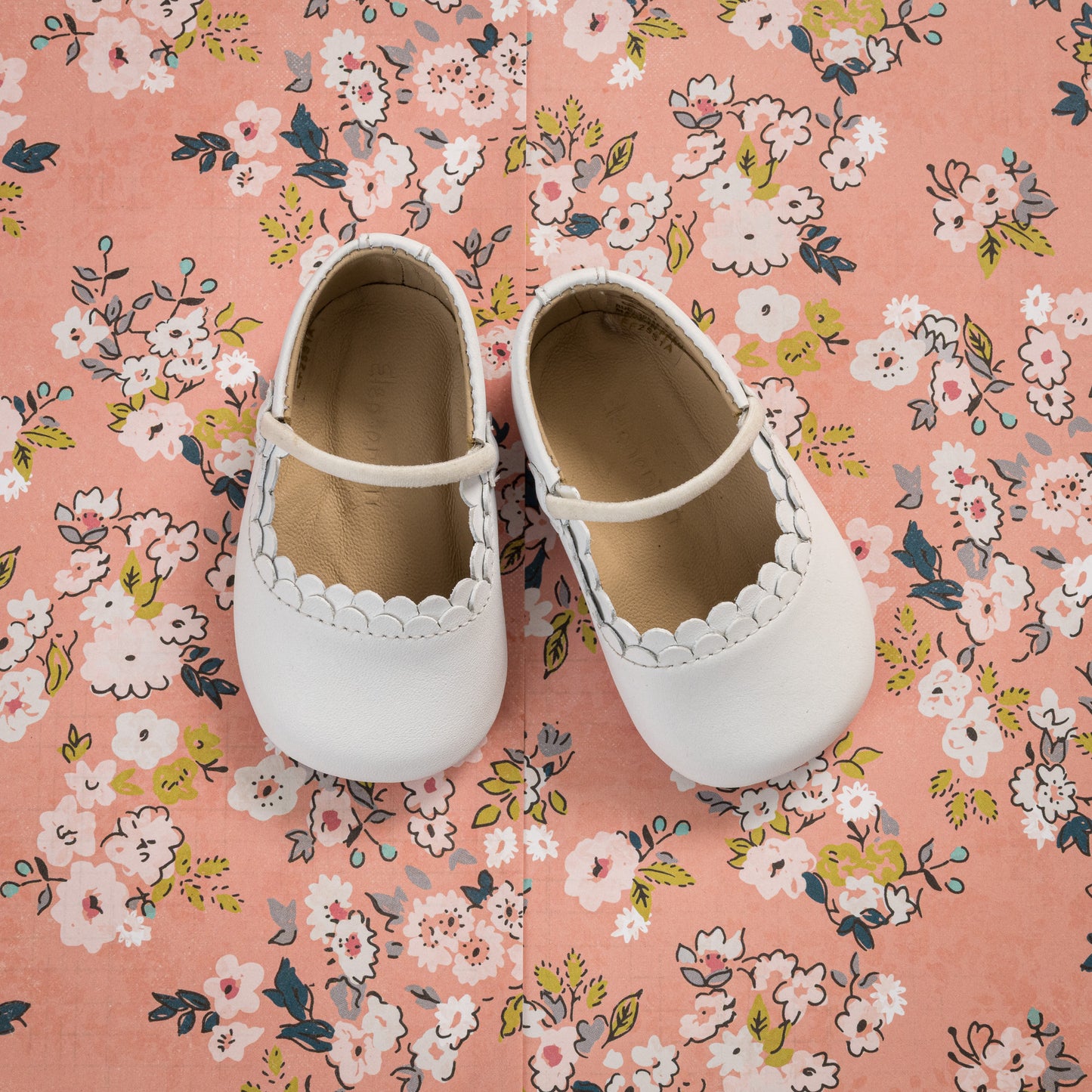 White baby ballet flats with scalloped edges and a single strap, on pink floral fabric background.
