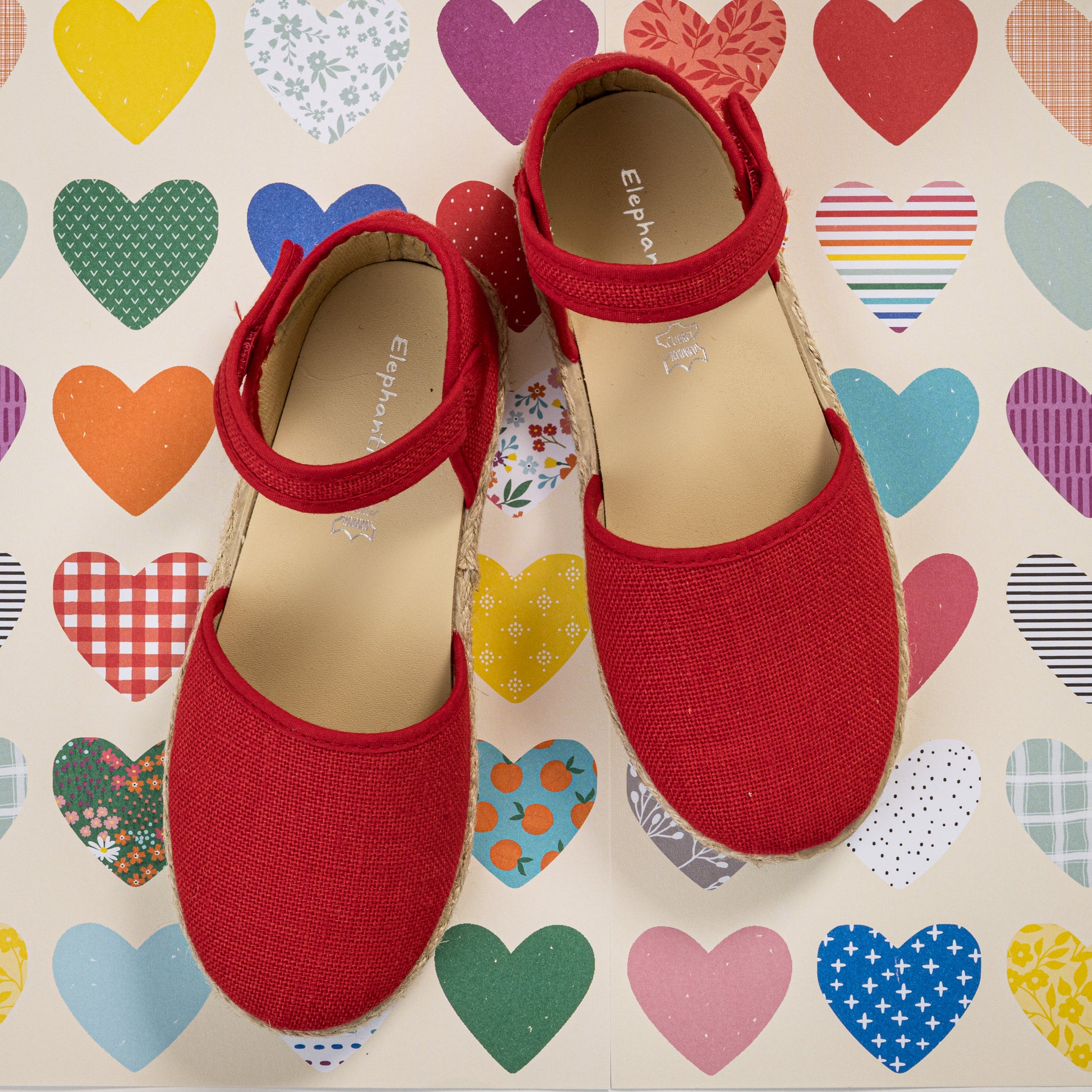 Red linen espadrille sandals for girls with adjustable ankle straps on colorful heart-patterned background.
