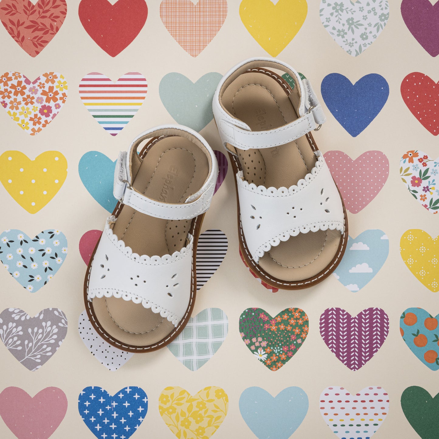 White toddler sandal with scalloped edges and brown sole on heart-patterned background.