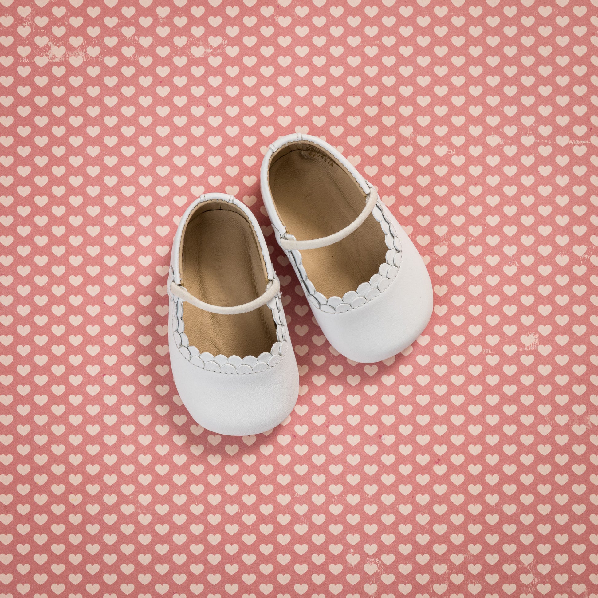 White baby Mary Jane shoes with scalloped edges and a strap, displayed on a pink heart-patterned background.