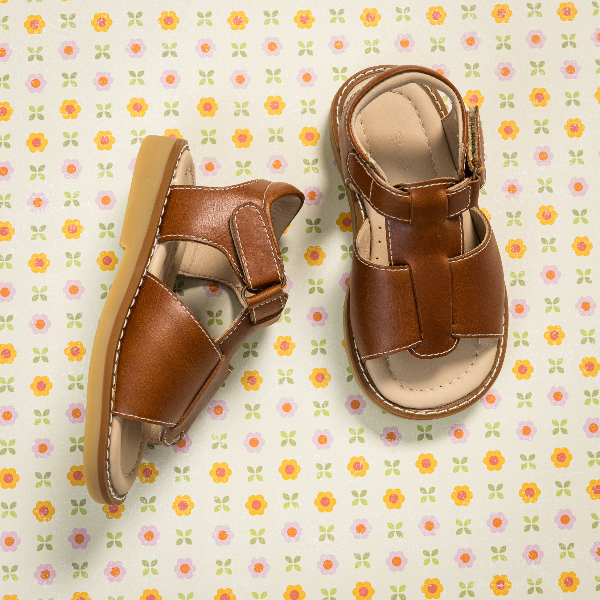 Natural brown leather boy sandal with open toe and velcro strap closure on patterned background.