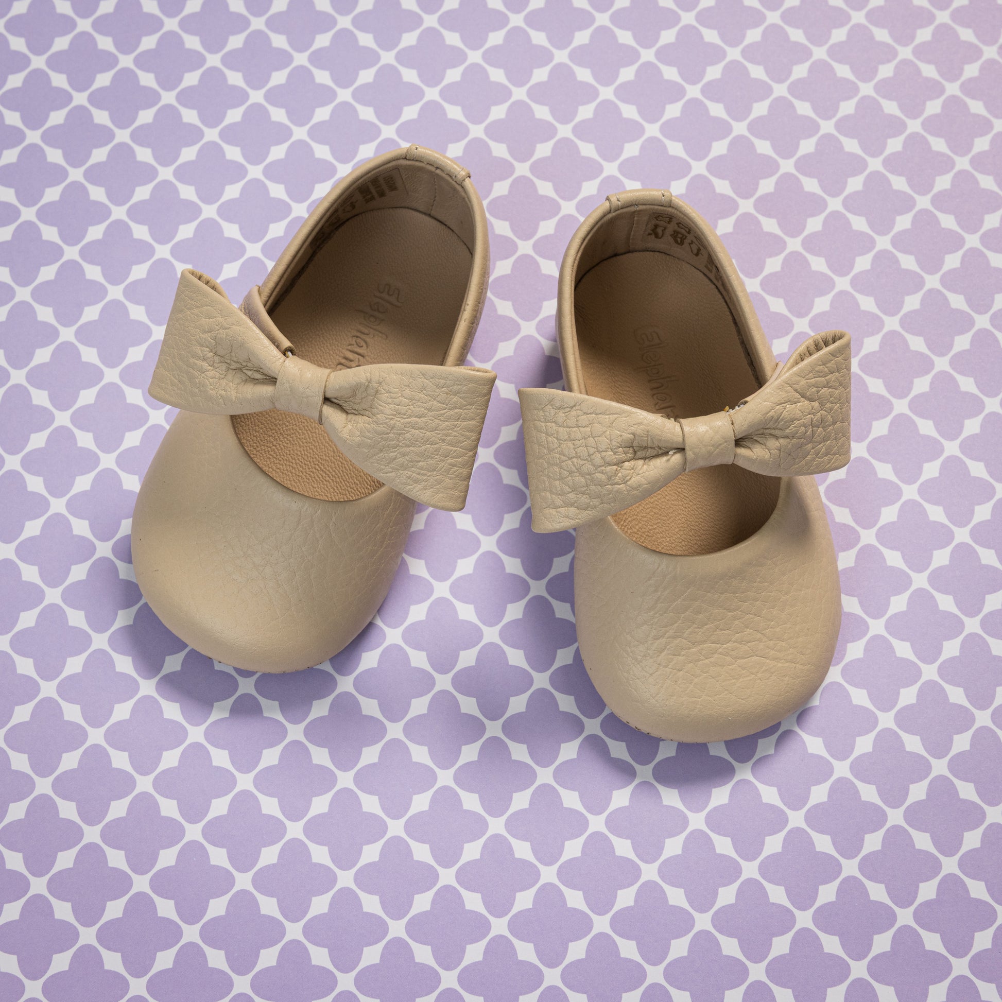 Cream-colored baby ballerina shoes with large textured bows and soft leather material on a patterned purple background.