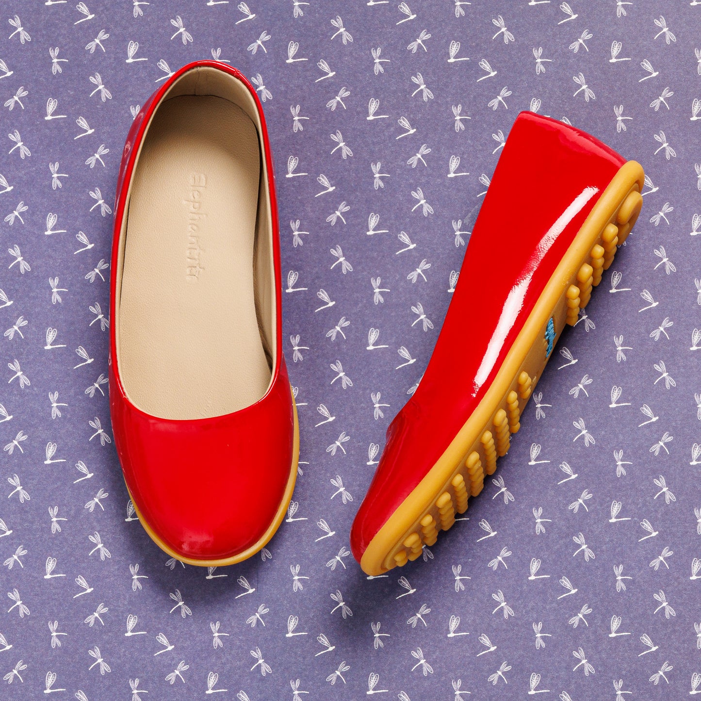 Shiny red patent leather ballerina flat with tan rubber sole for toddlers on purple patterned background.