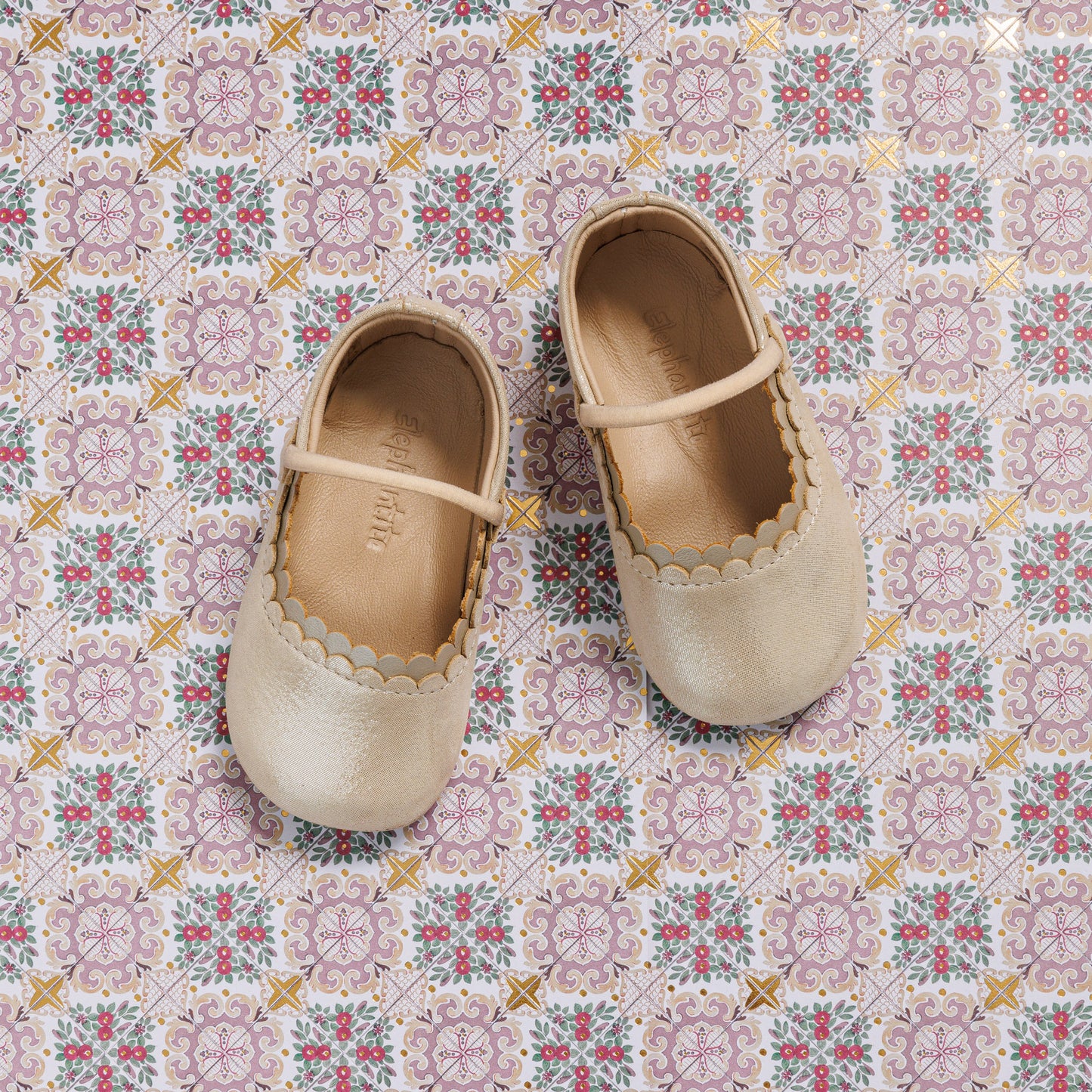 Cream-colored baby ballet flats with scalloped edges and a thin strap across the top, displayed on a patterned tile.