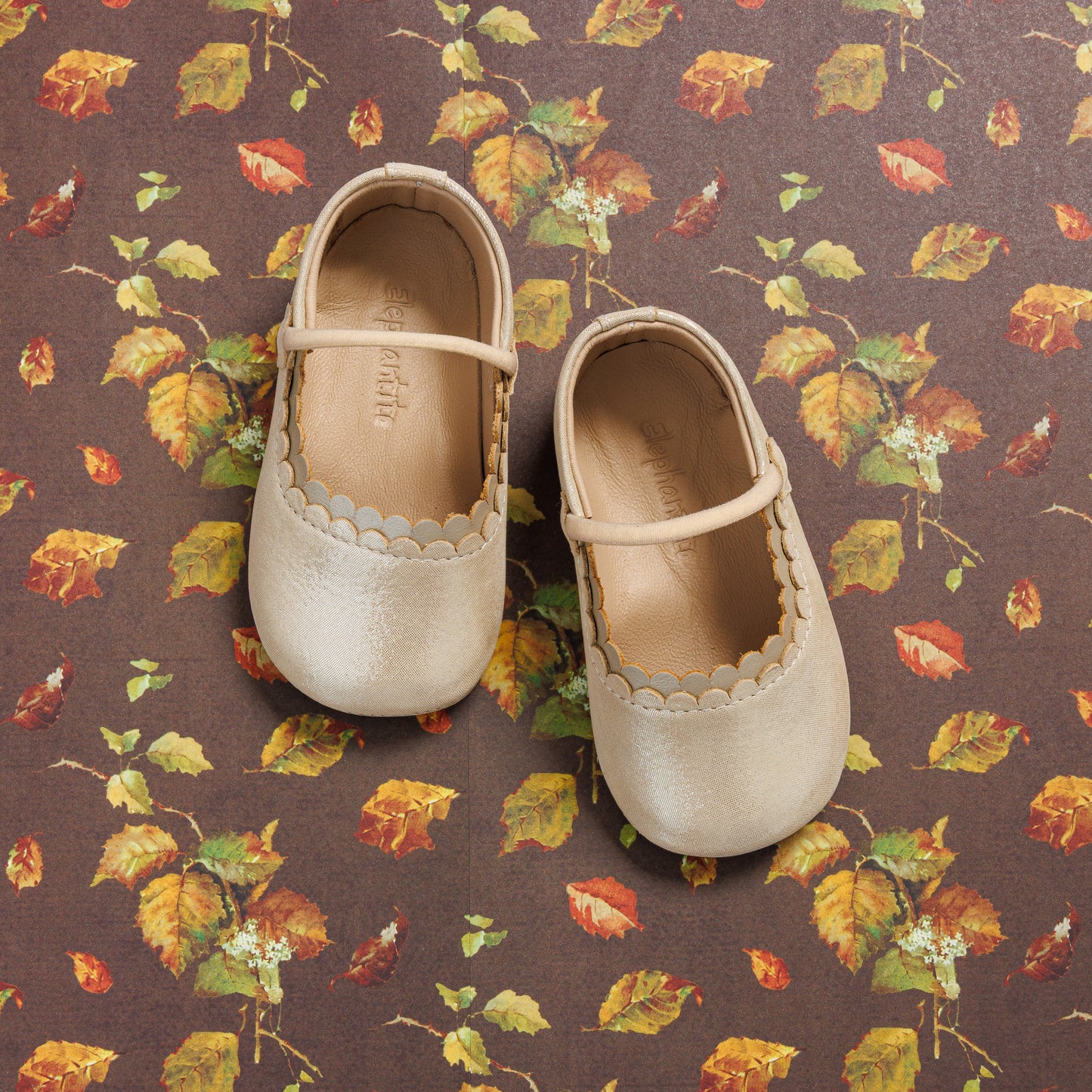 Cream-colored baby ballet shoes with scalloped edges and soft leather on a floral fabric background.