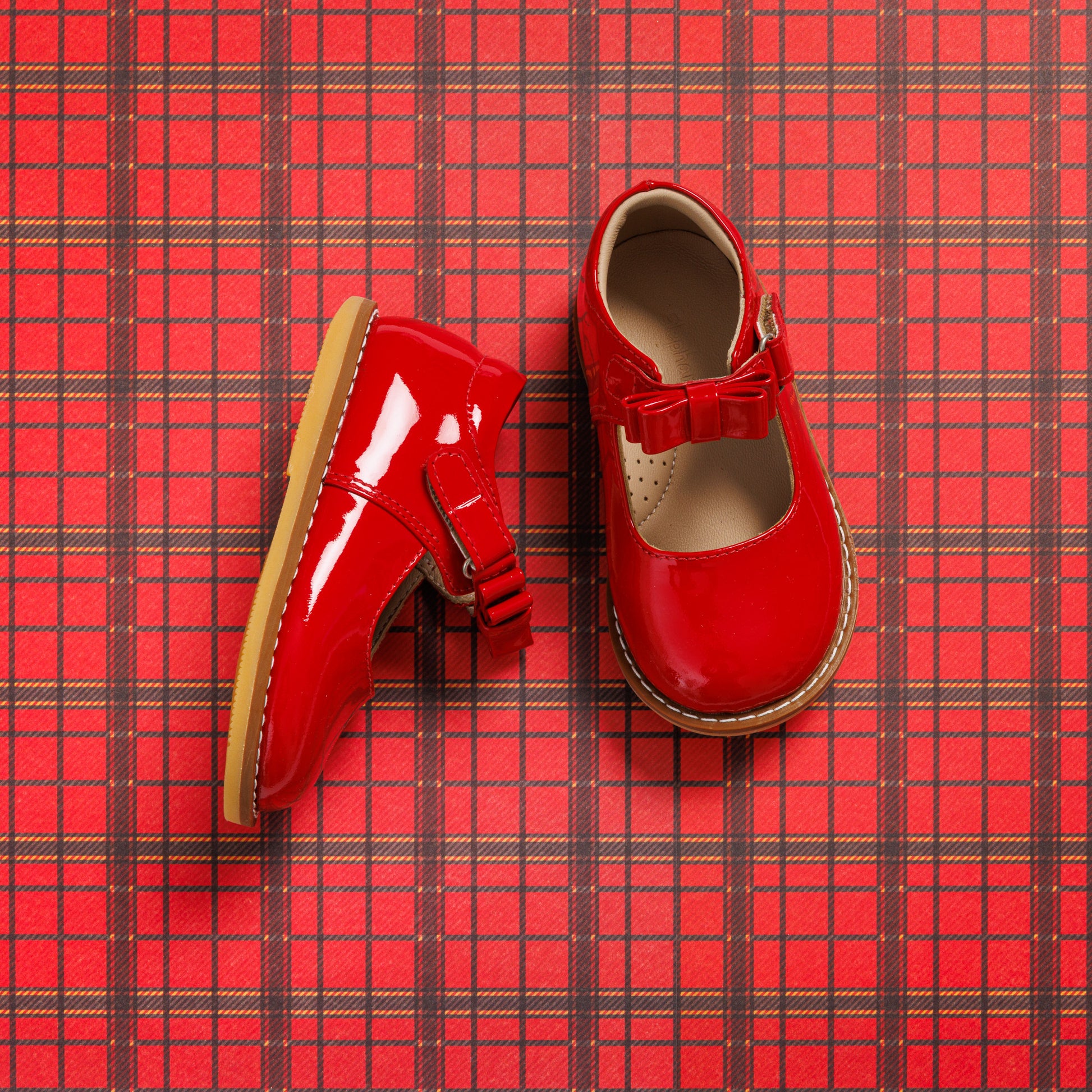 Glossy red toddler Mary Jane shoes with a bow detail and beige insoles on red plaid background.