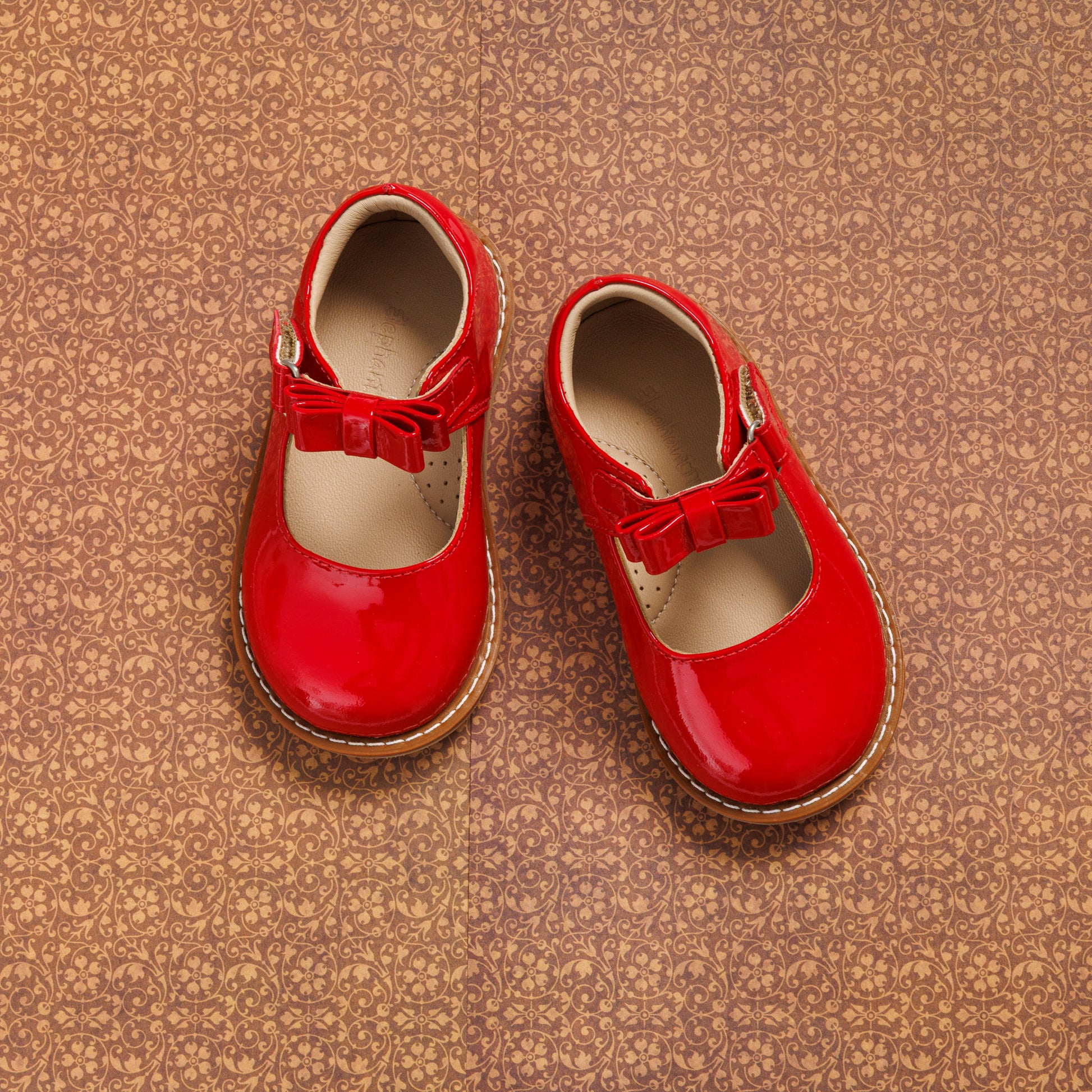 Glossy red toddler Mary Jane shoes with a bow detail and brown soles on a textured background.