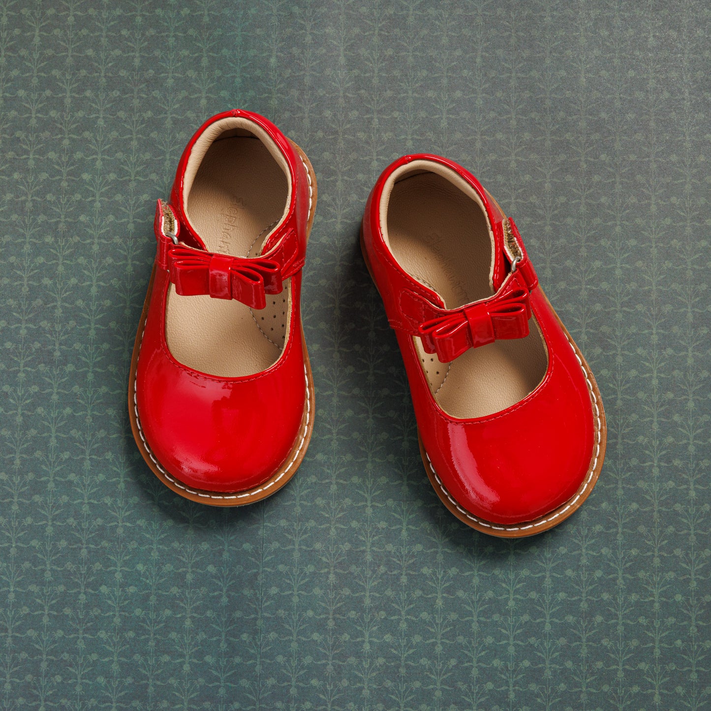 Shiny red toddler Mary Jane shoes with a bow detail and tan sole on green patterned background.