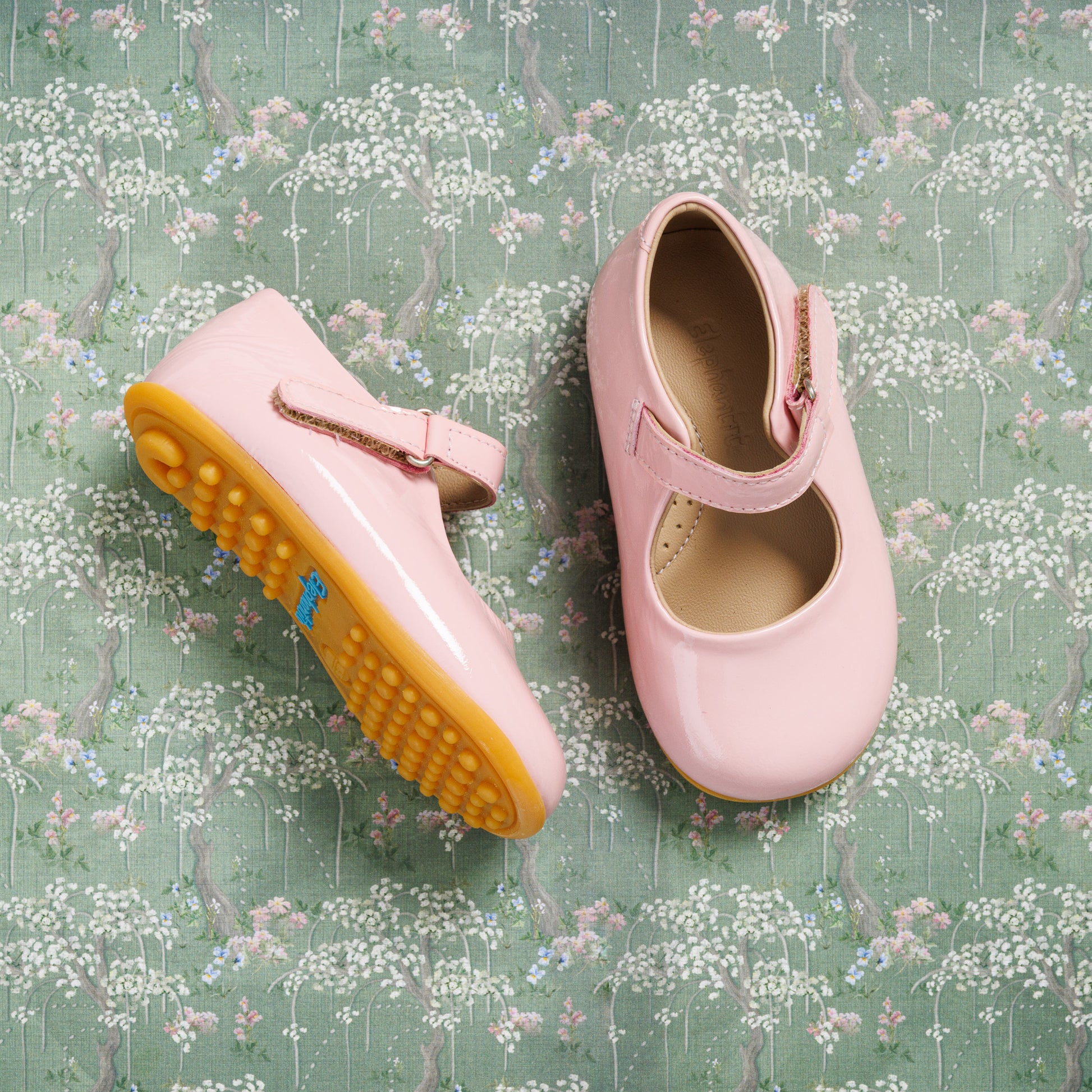 Pink toddler Mary Jane shoes with a rounded toe and yellow flexible sole on a floral background.