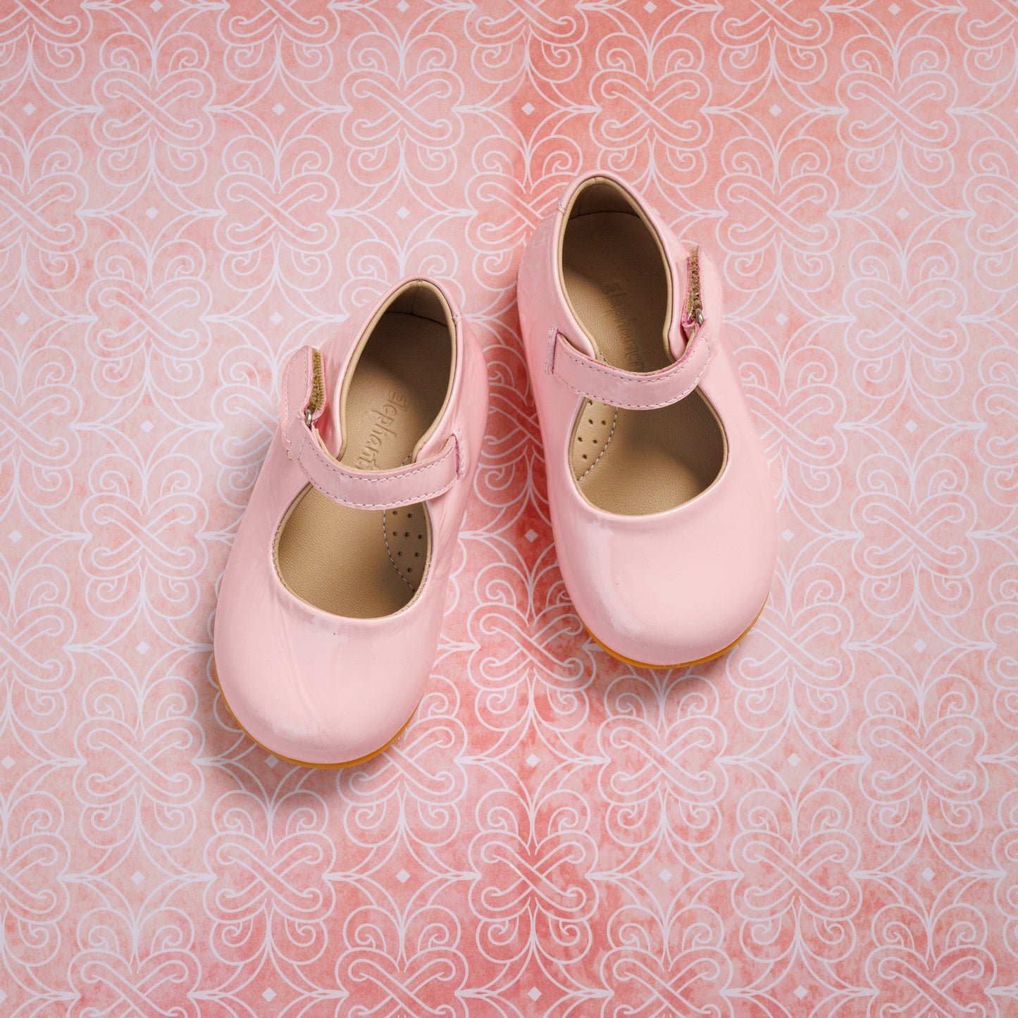 Light pink toddler Mary Jane shoes with a velcro strap and tan soles on a patterned background.