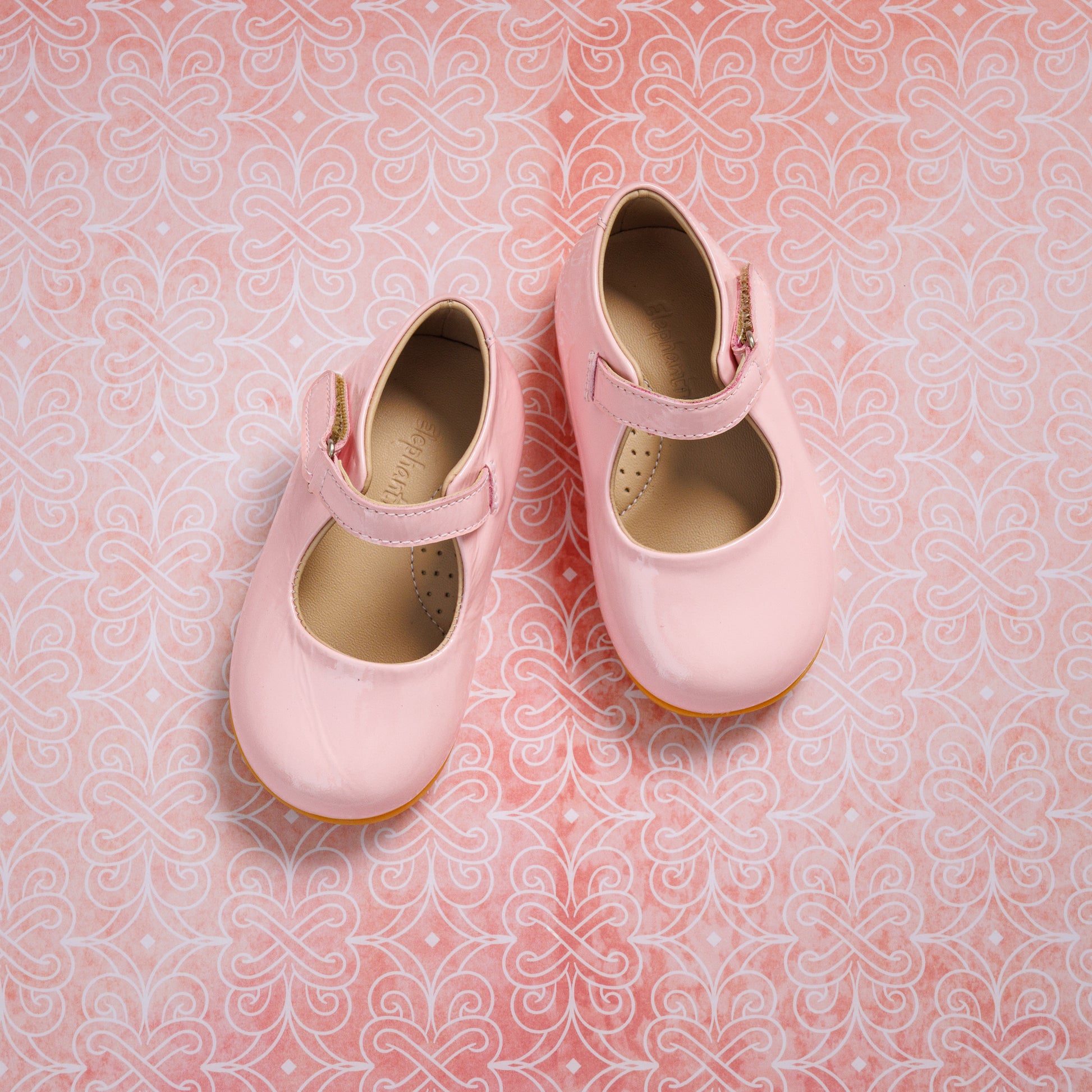 Light pink toddler Mary Jane shoes with a velcro strap and tan soles on a patterned background.
