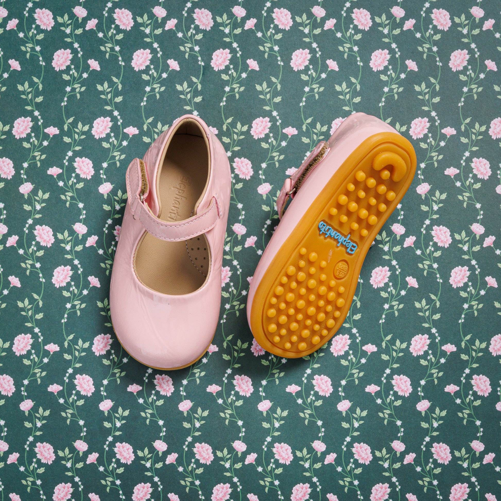 Toddler pink leather Mary Jane shoes with adjustable strap and textured mustard sole on floral background.