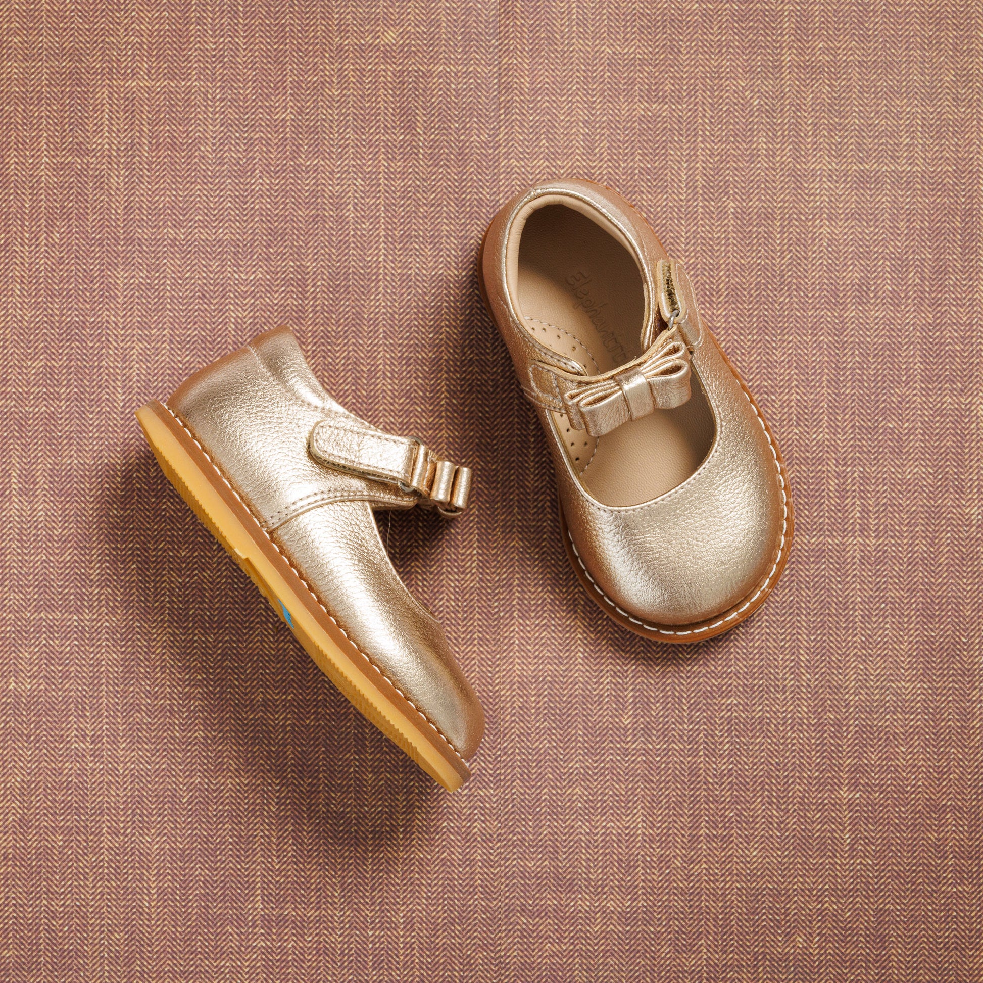 Gold toddler Mary Jane shoes with bow detail and tan rubber soles on textured fabric background.