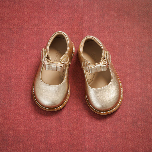 Gold toddler Mary Jane shoes with a small bow and buckle strap on a red textured background.