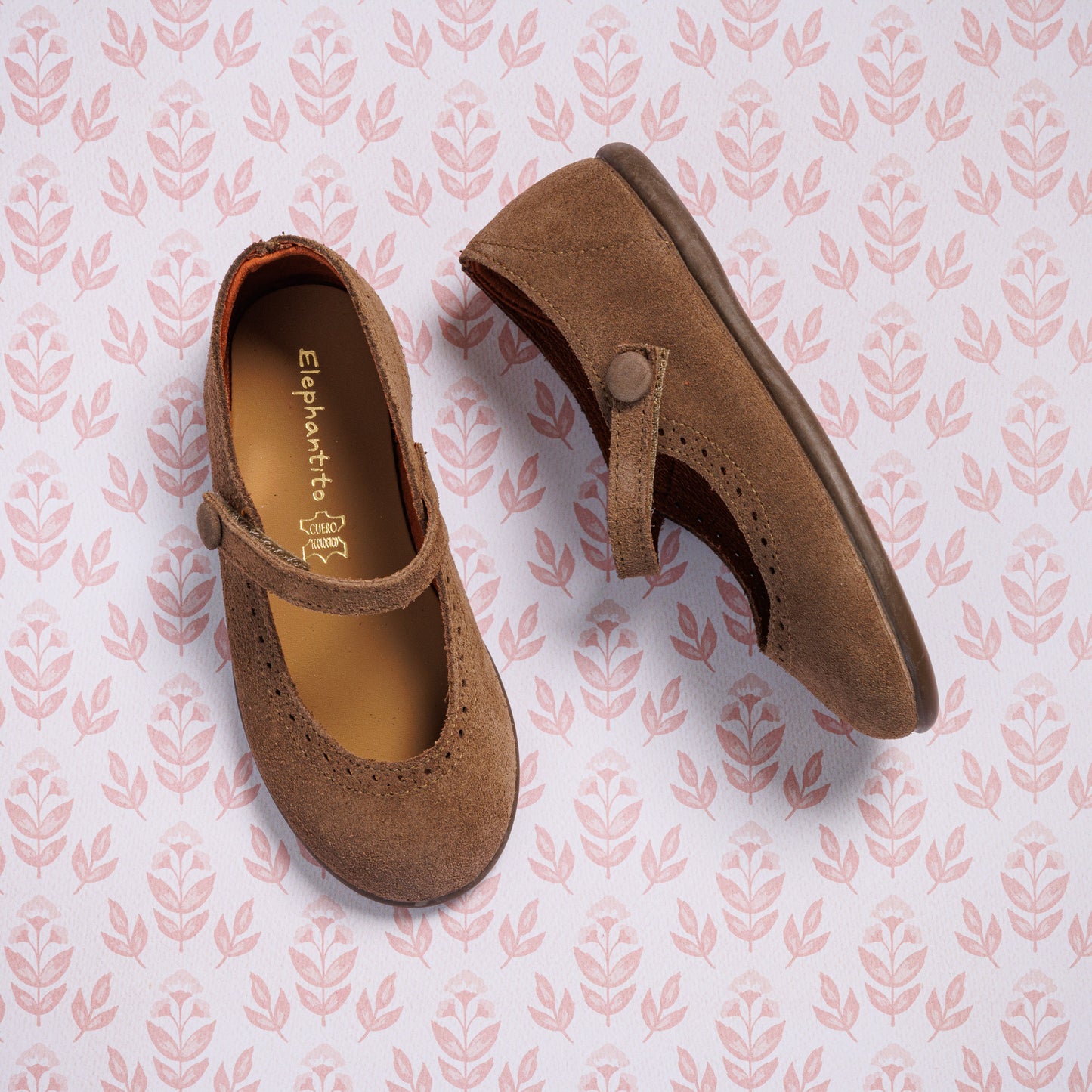 The Elephantito Suede Mary Jane Beige shoes for girls, featuring a button strap, are displayed on a pink background with a repeating leaf pattern.