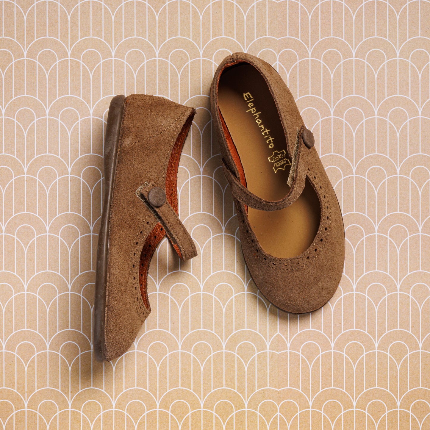 A pair of Elephantito Suede Mary Jane Beige shoes for girls, with one flat and one on its side, shown on a tan geometric patterned background.