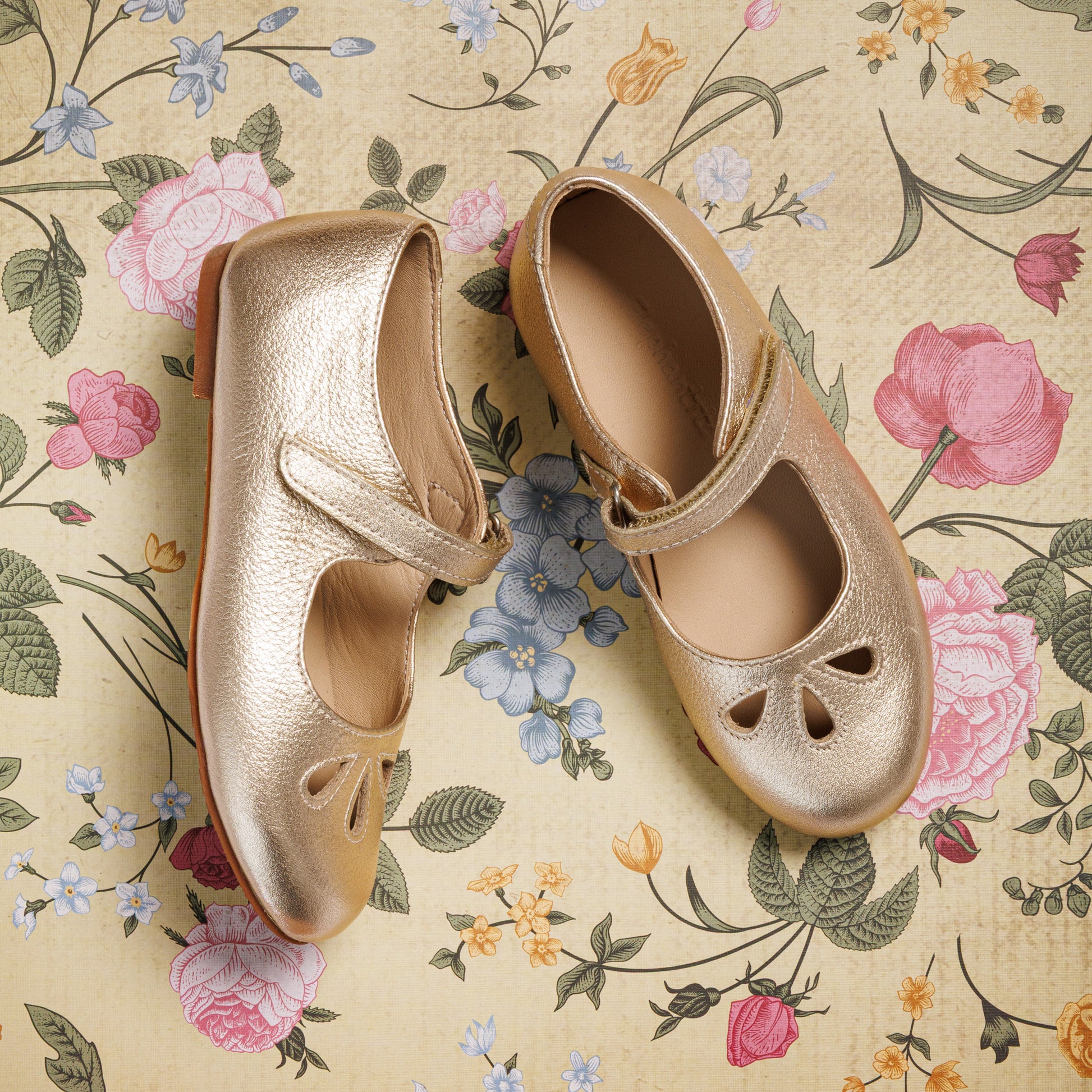 Metallic gold toddler Mary Jane shoes with cutout heart details and Velcro strap closure on floral background.