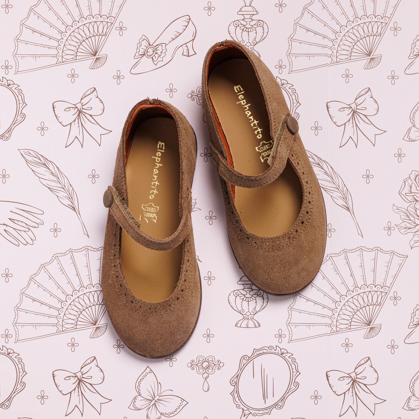 A pair of Suede Mary Jane Beige shoes by Elephantito with buttoned straps is displayed on paper featuring illustrated fans, bows, mirrors, feathers, and perfume bottles.