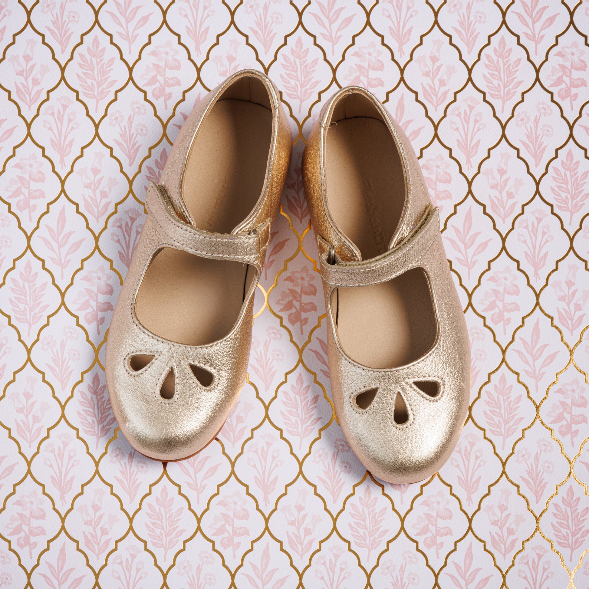 Gold toddler Mary Jane shoes with cut-out details and a Velcro strap on patterned background.