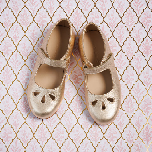 Gold toddler Mary Jane shoes with cut-out details and a Velcro strap on patterned background.