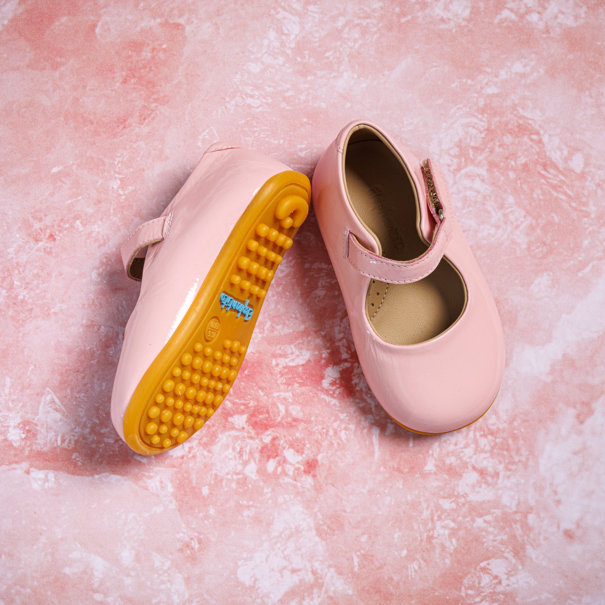 Pink toddler Mary Jane shoes with adjustable strap and textured yellow sole on pink background.