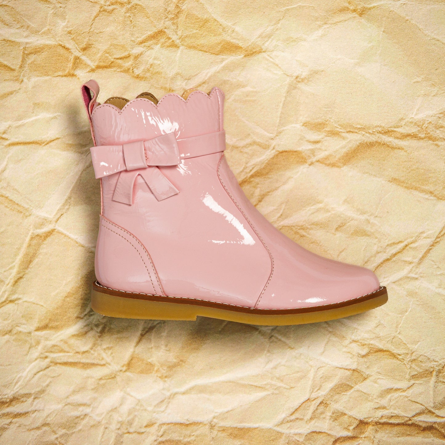 Glossy light pink toddler bootie with scalloped top edge and decorative bow, tan sole.