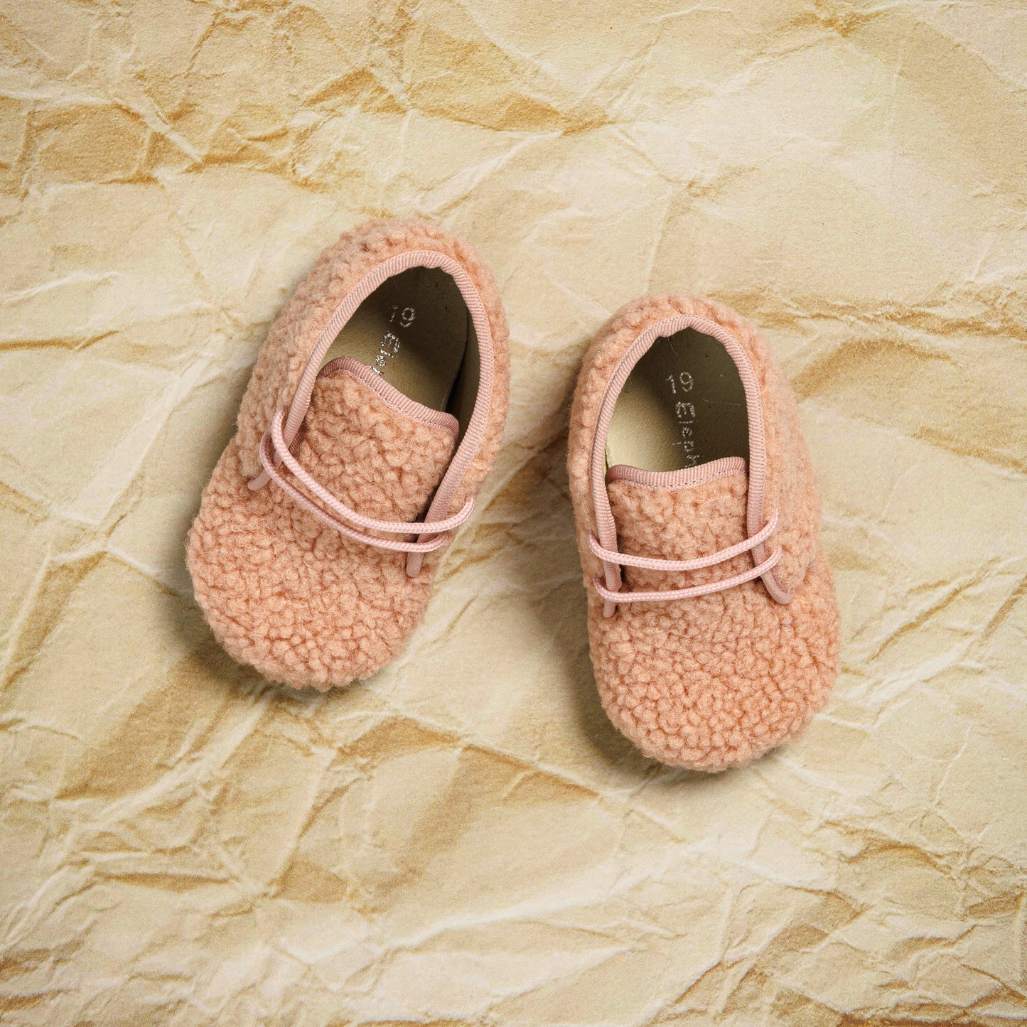 Pink shearling baby booties with soft textured fabric and light pink laces on crinkled beige paper background.