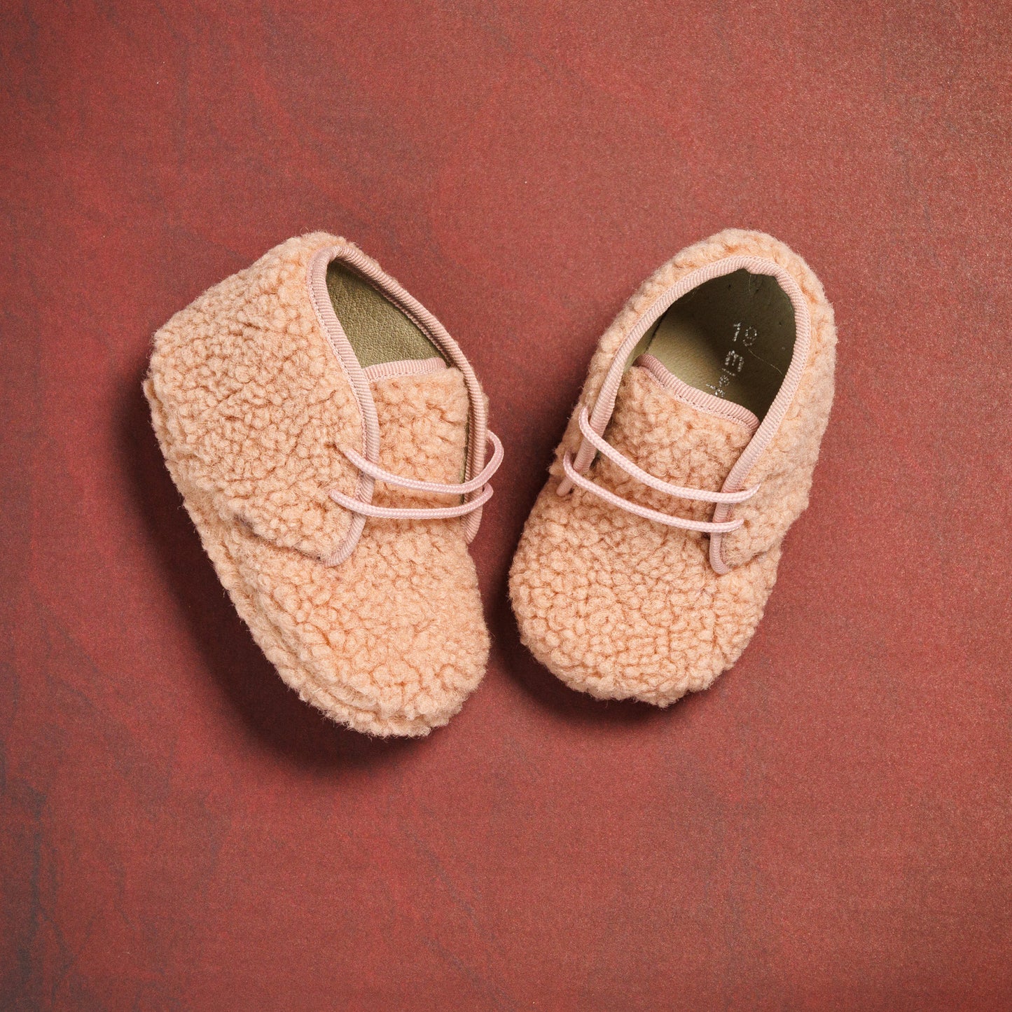 Pink shearling baby booties with soft textured exterior and elastic laces on red background.