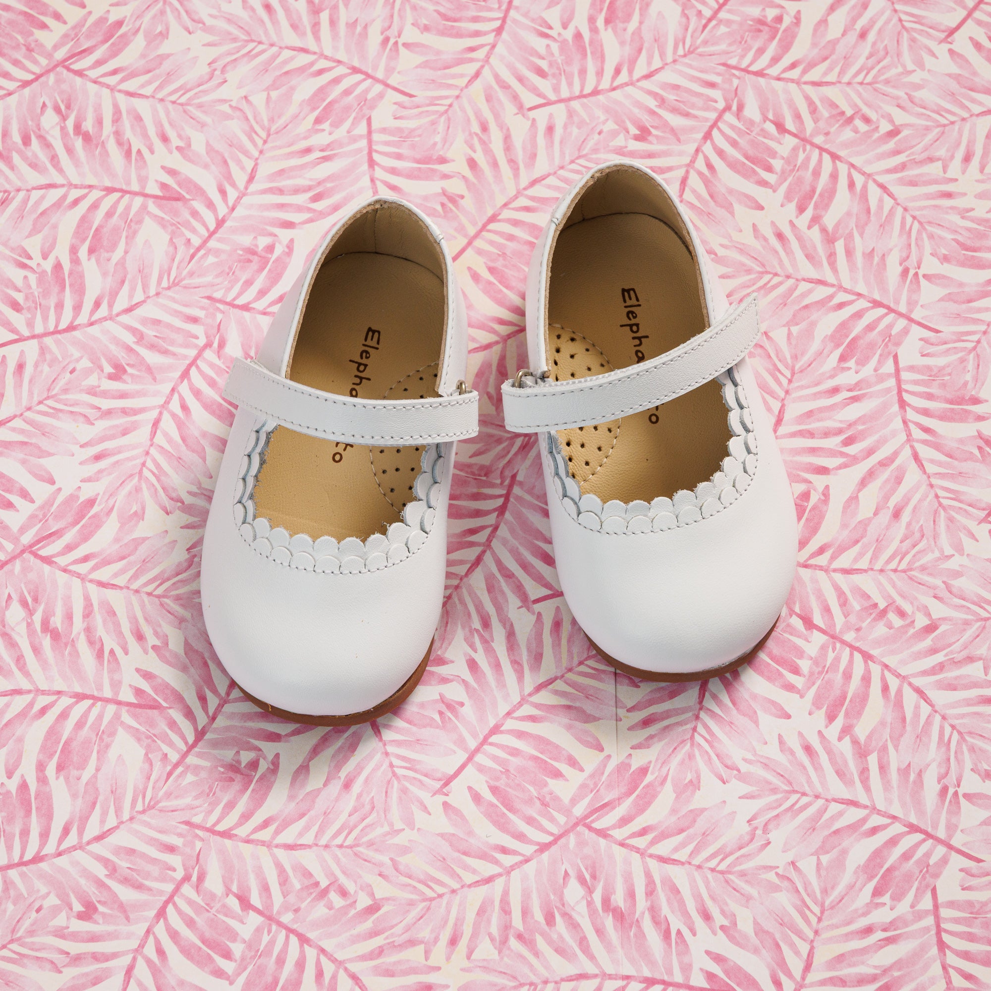 Elephantito - White toddler Mary Jane shoes with scalloped edges and a single strap, set on pink textured background.