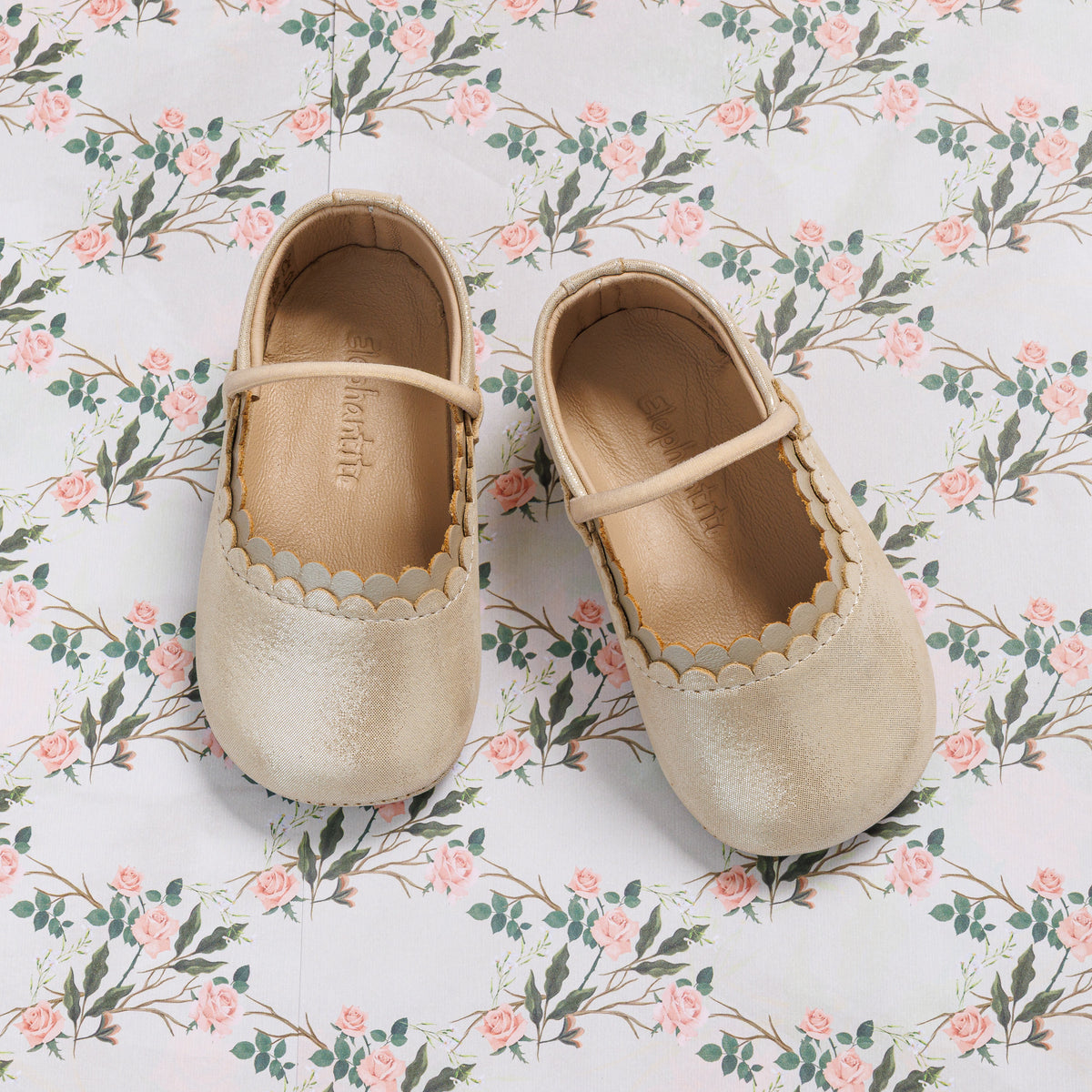 Soft gold baby ballet shoes with scalloped edges and delicate strap on floral background.