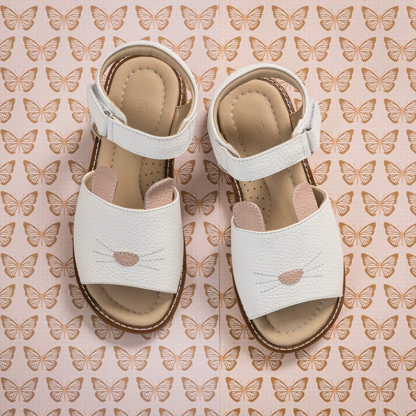 White toddler sandals with bunny face design and adjustable ankle straps on butterfly-patterned background.