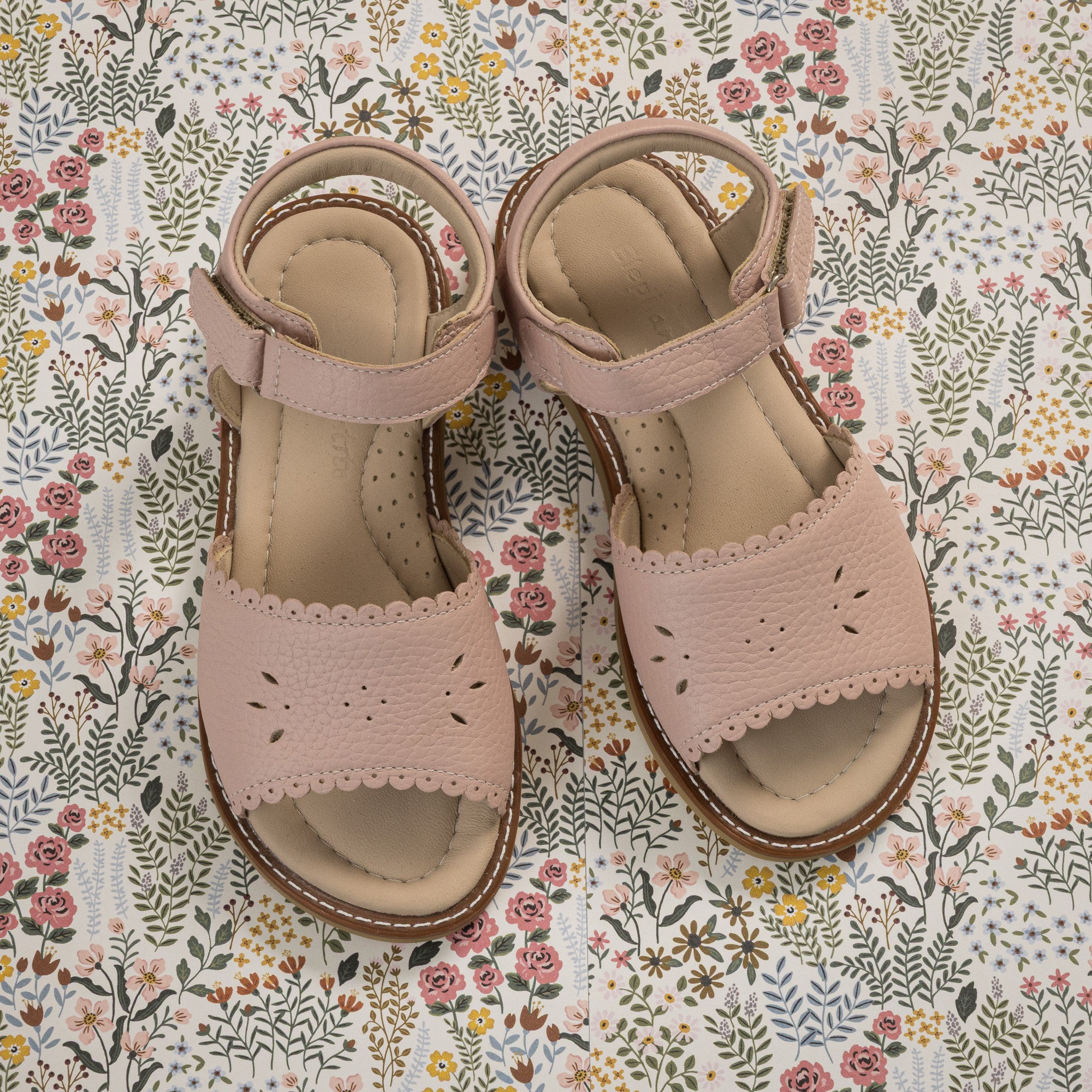 Pink toddler sandals with scalloped edges, perforated floral design, and ankle straps on floral background.