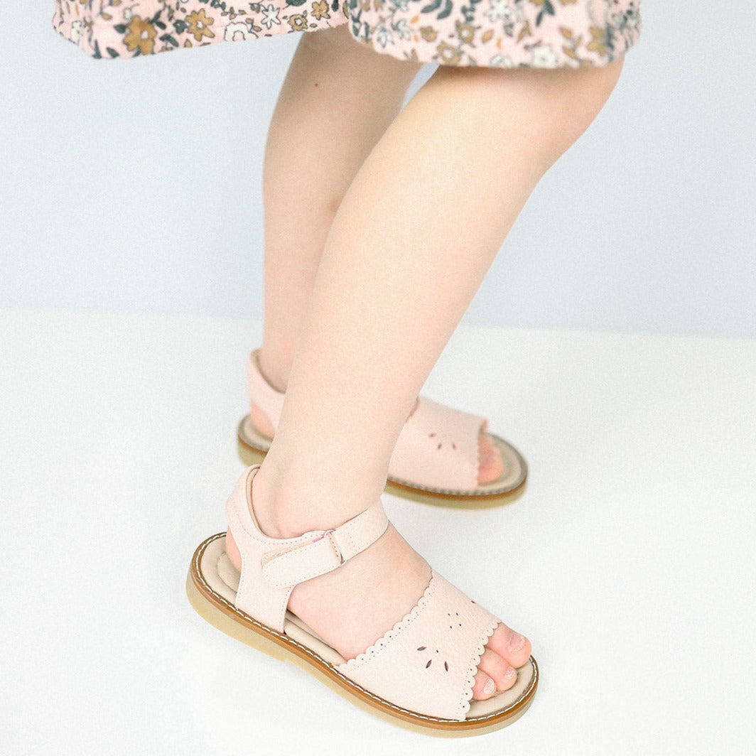 Pink toddler sandal with scalloped edges and ankle strap on child’s feet.