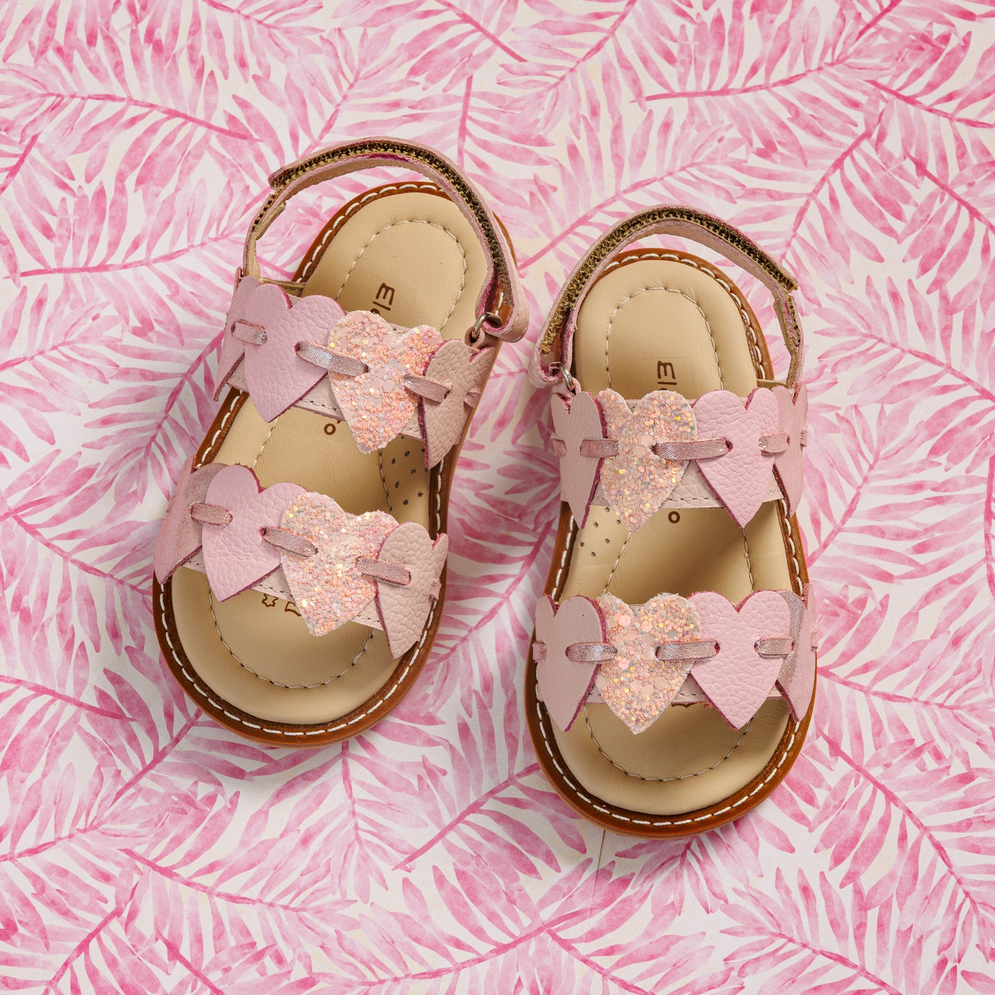 Elephantito - Pink toddler sandals with heart-shaped straps and cushioned tan insoles on a pink leaf-patterned background.