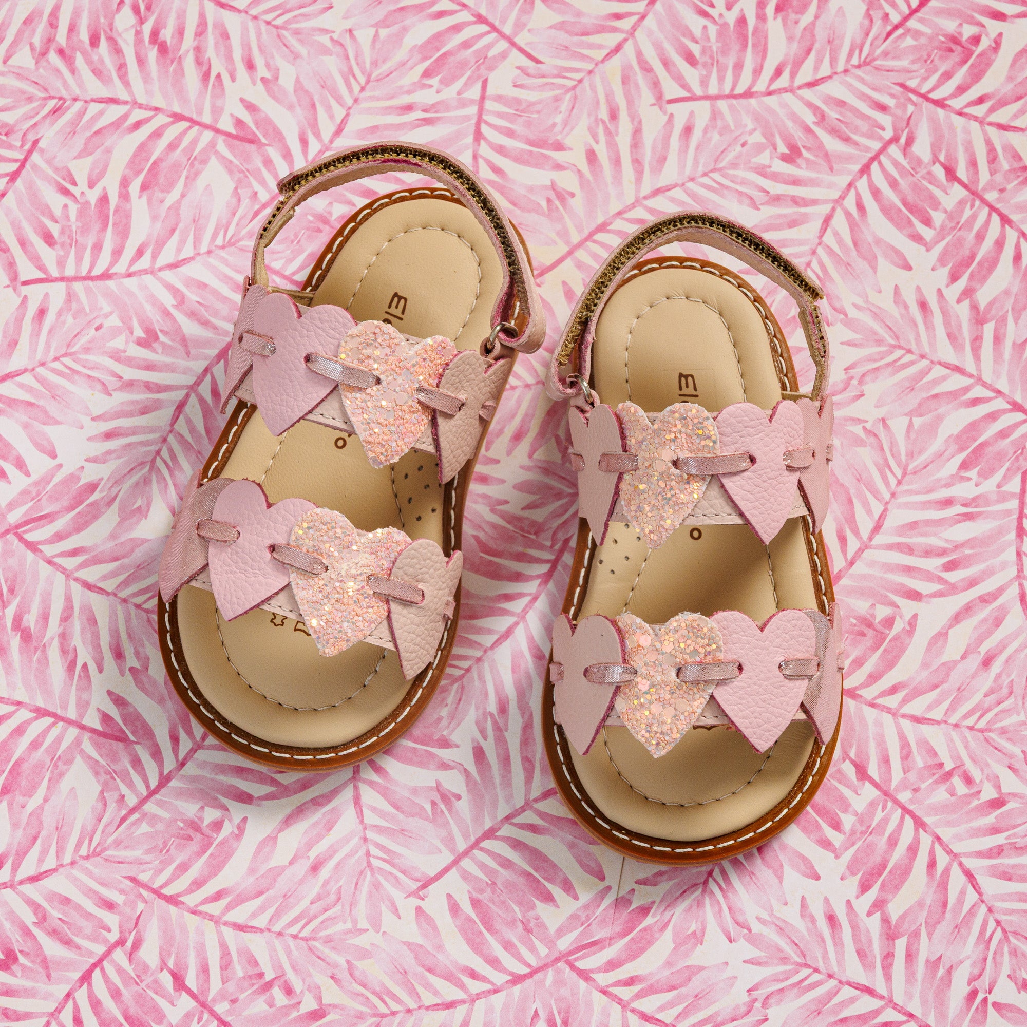 Elephantito - Pink toddler sandals with heart-shaped straps and cushioned tan insoles on a pink leaf-patterned background.