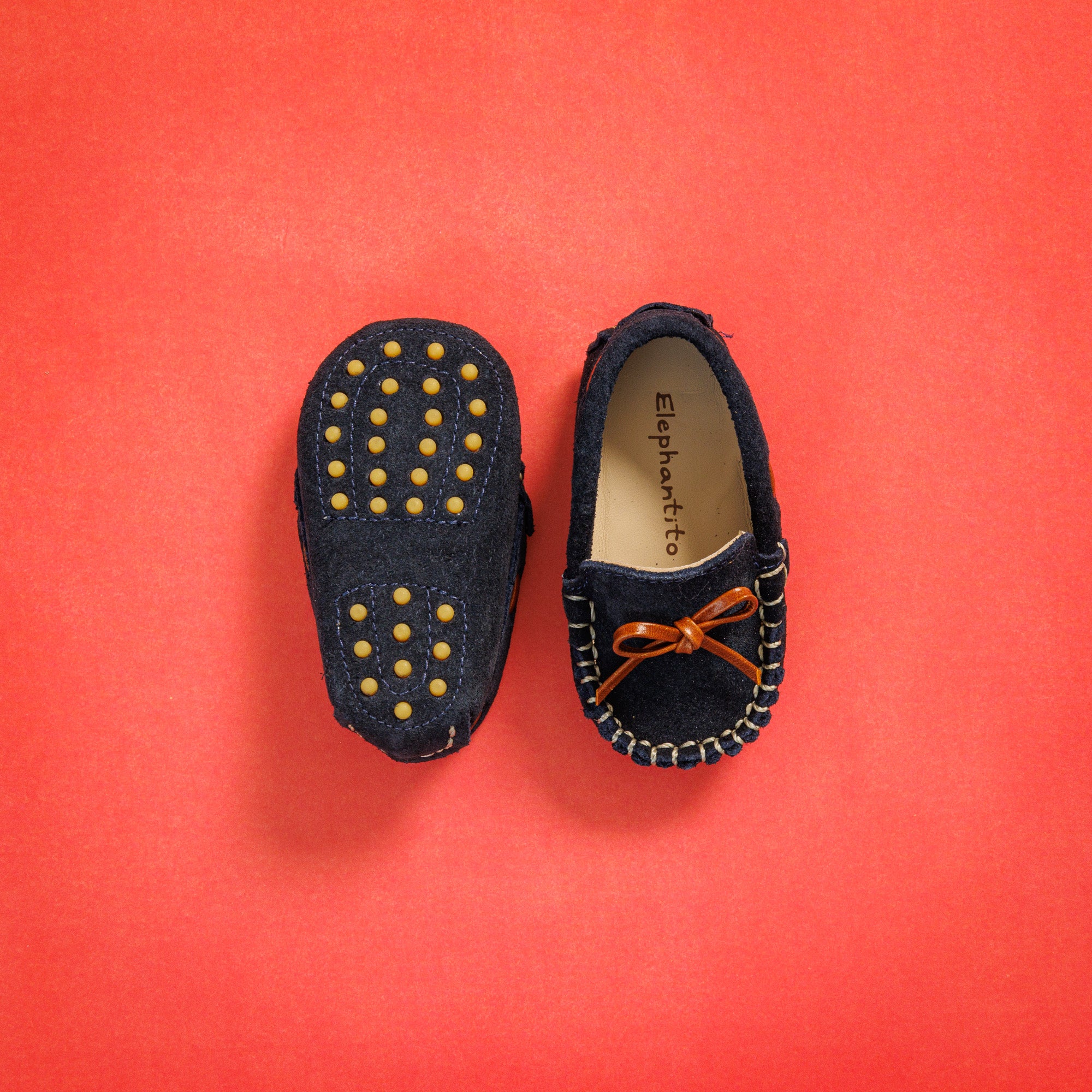 Elephantito - Navy blue baby suede driver loafers with brown bow and rubber grip soles on red background.